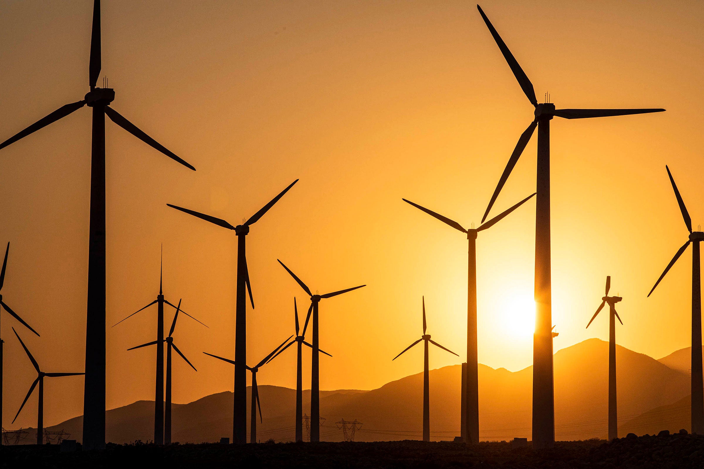 Several wind turbines stand against an orange sky. A bright yellow sun hangs low over mountains in the background.