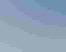 a person stands on a beach and looks through a spotting scope, facing left. In the background is the ocean and a blue sky with sparse clouds.