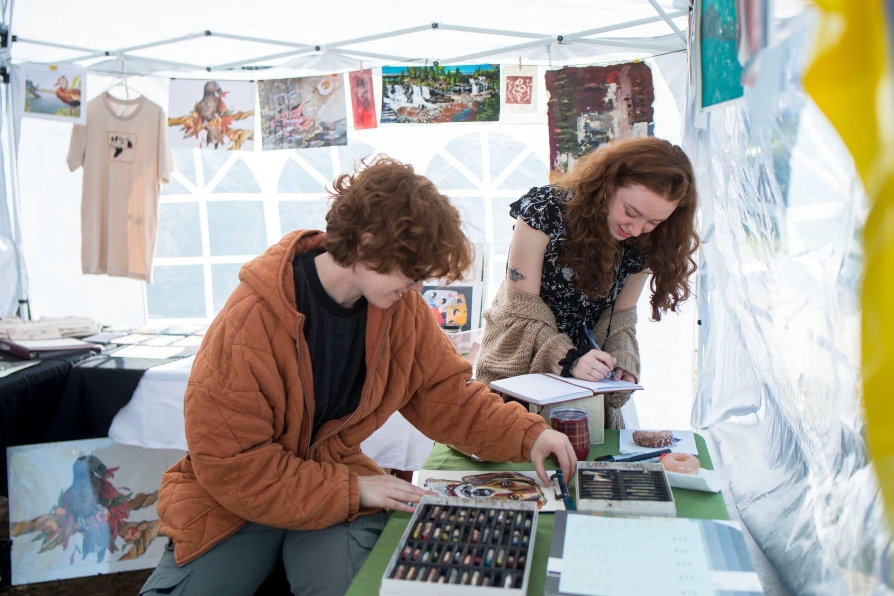 Two siblings with red hair prepare their art to sell at an outdoor stall
