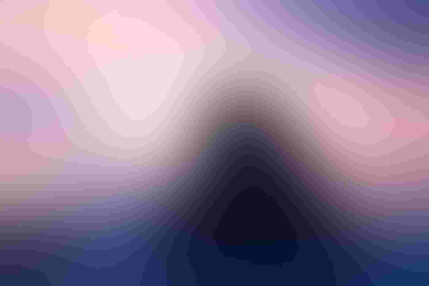 A giant, peaked rock surrounded by water is centered in the frame, backlit and silhouetted, blue waves crashing at its base. Two tiny gulls sit atop the rock against a pink and purple sky.