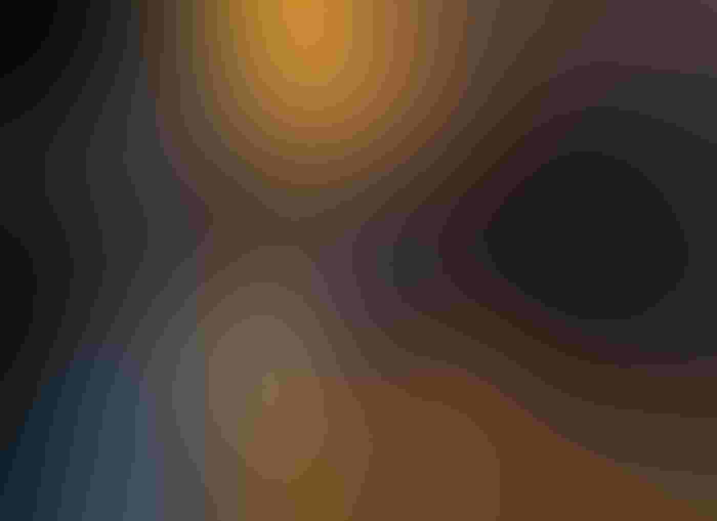 A male Wood Duck with a green head, chestnut breast, and red bill glides toward the camera over smooth water. Photographed at eye-level, the bird is in the center of the mostly-dark frame. The water reflects a blurred pop of orange in the background.