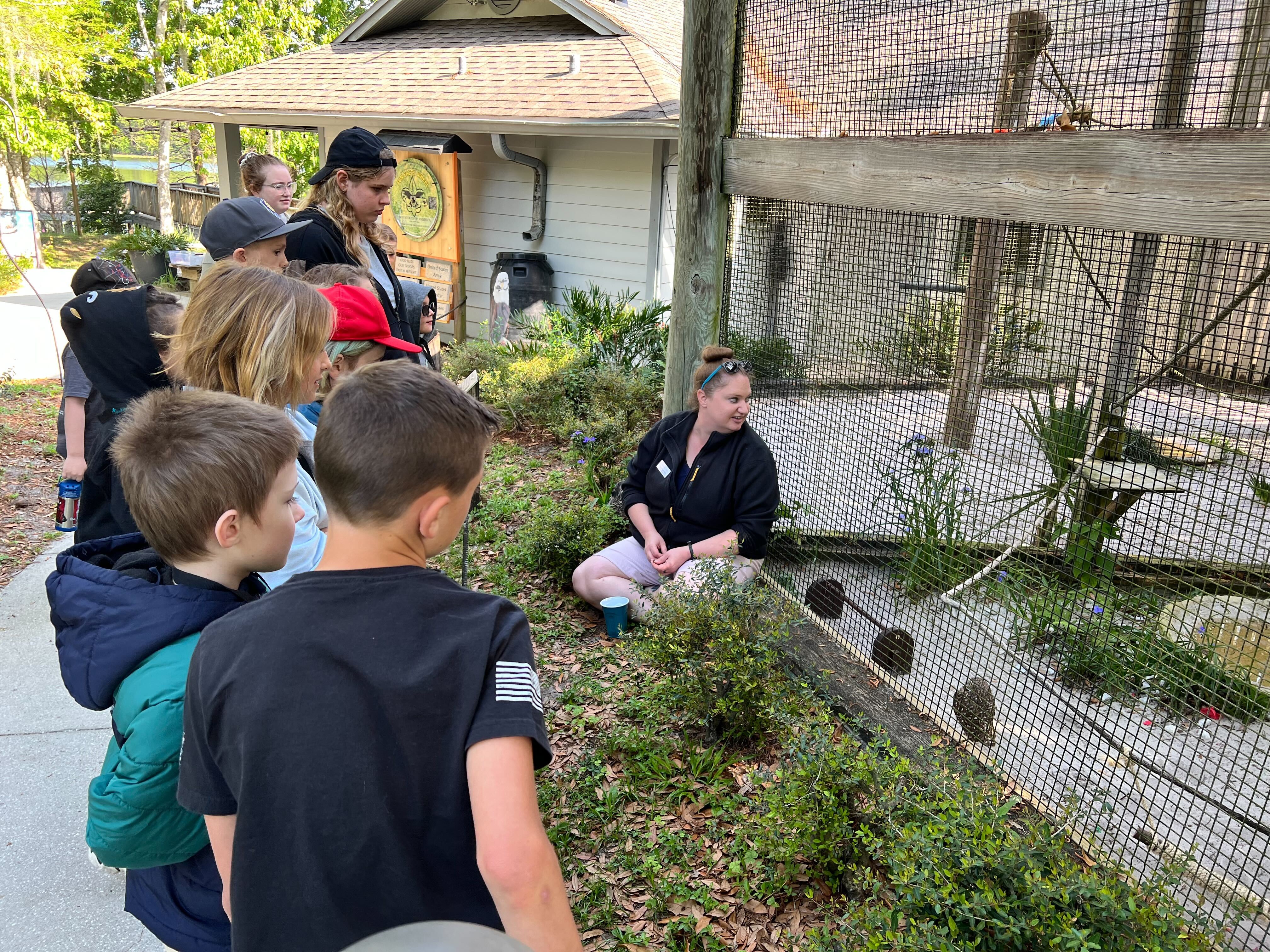 A woman sits in front of a bird enclosure and gestures toward a Burrowing Owl while kids look on.