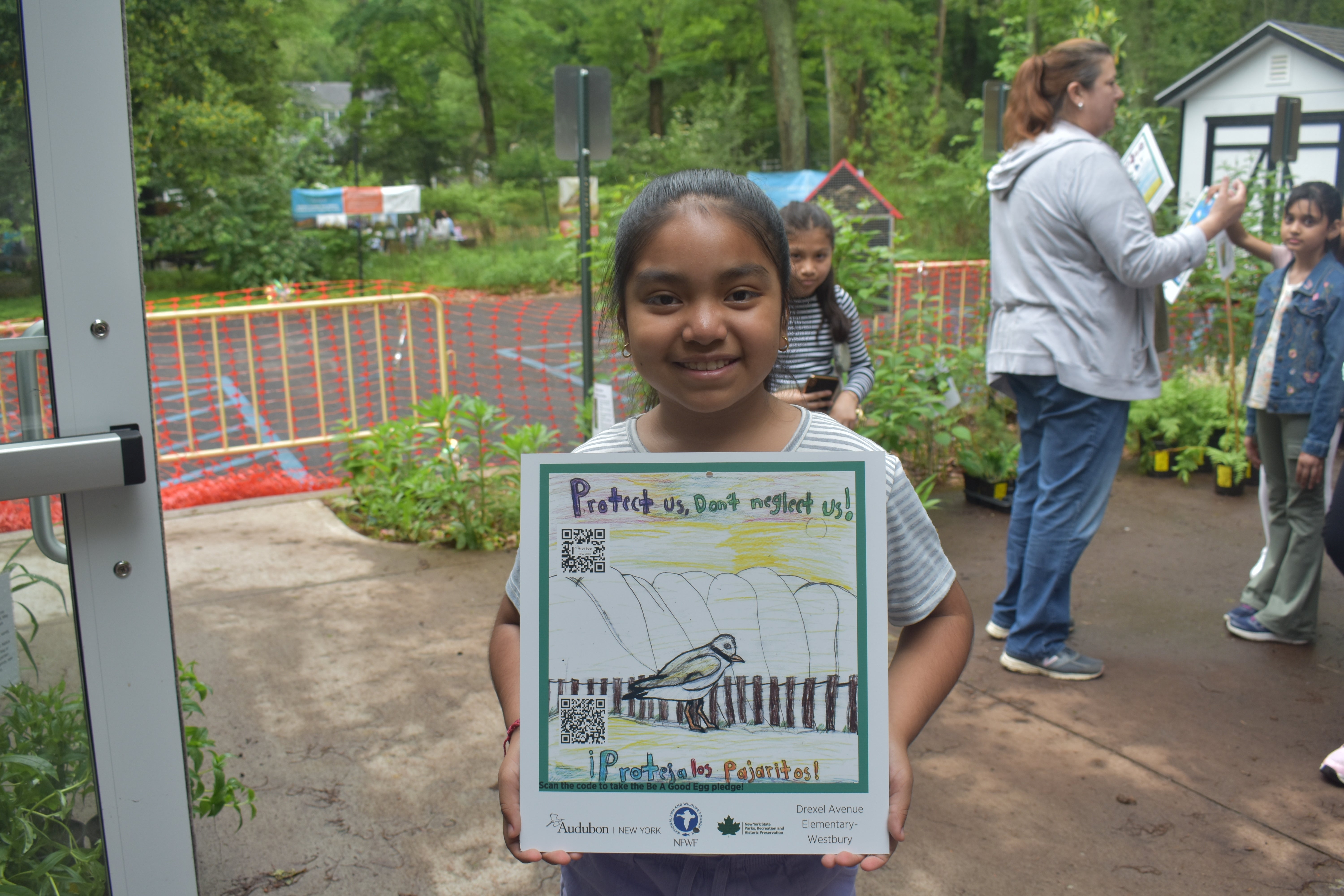 A child smiles as she holds up her shorebird sign, featuring a colorful illustration of a Piping Plover and hand-written text that reads: Protect us, don't neglect us! Proteja los pajaritos!