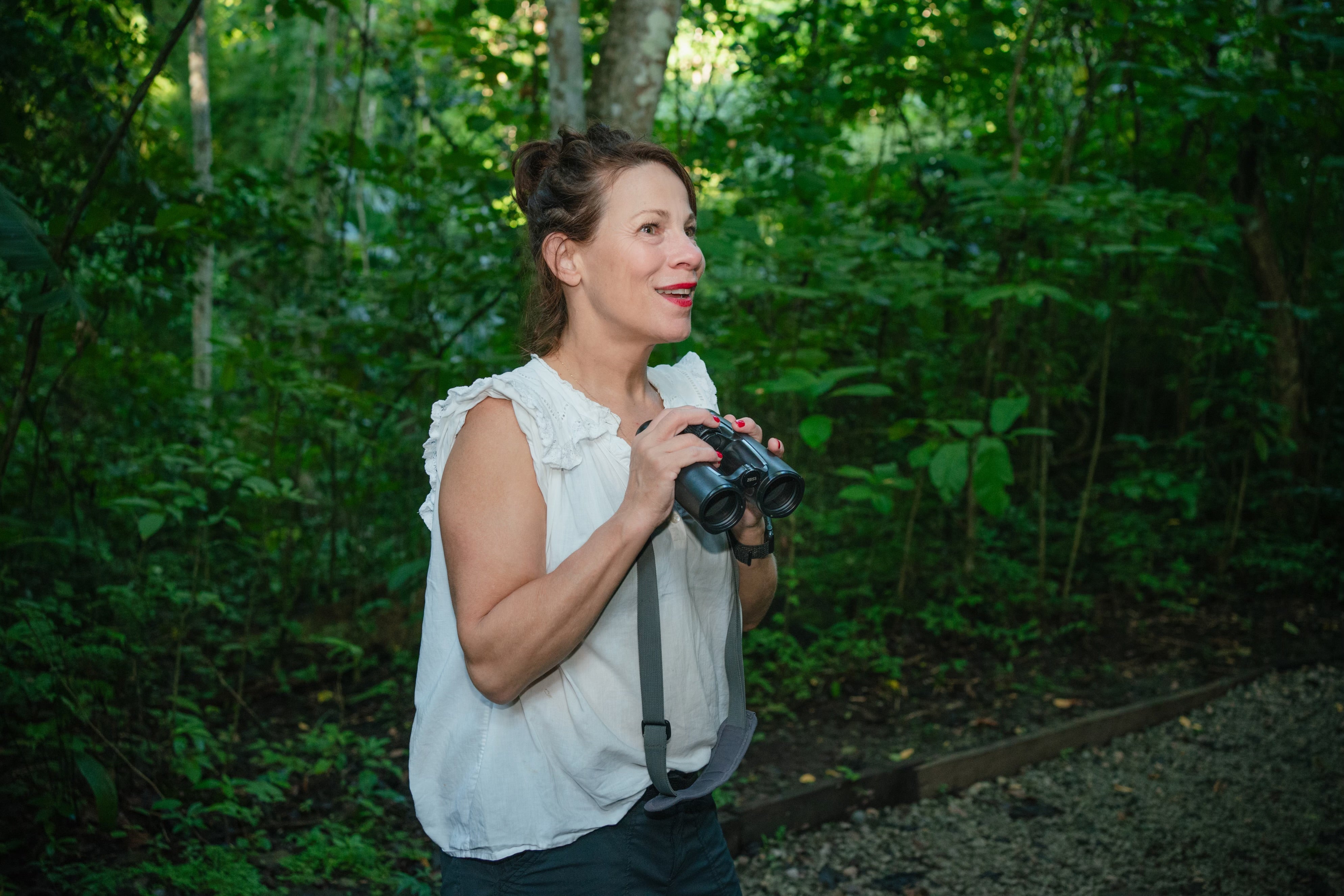 Lili Taylor stands in a lush, green forest holding binoculars and smiling.