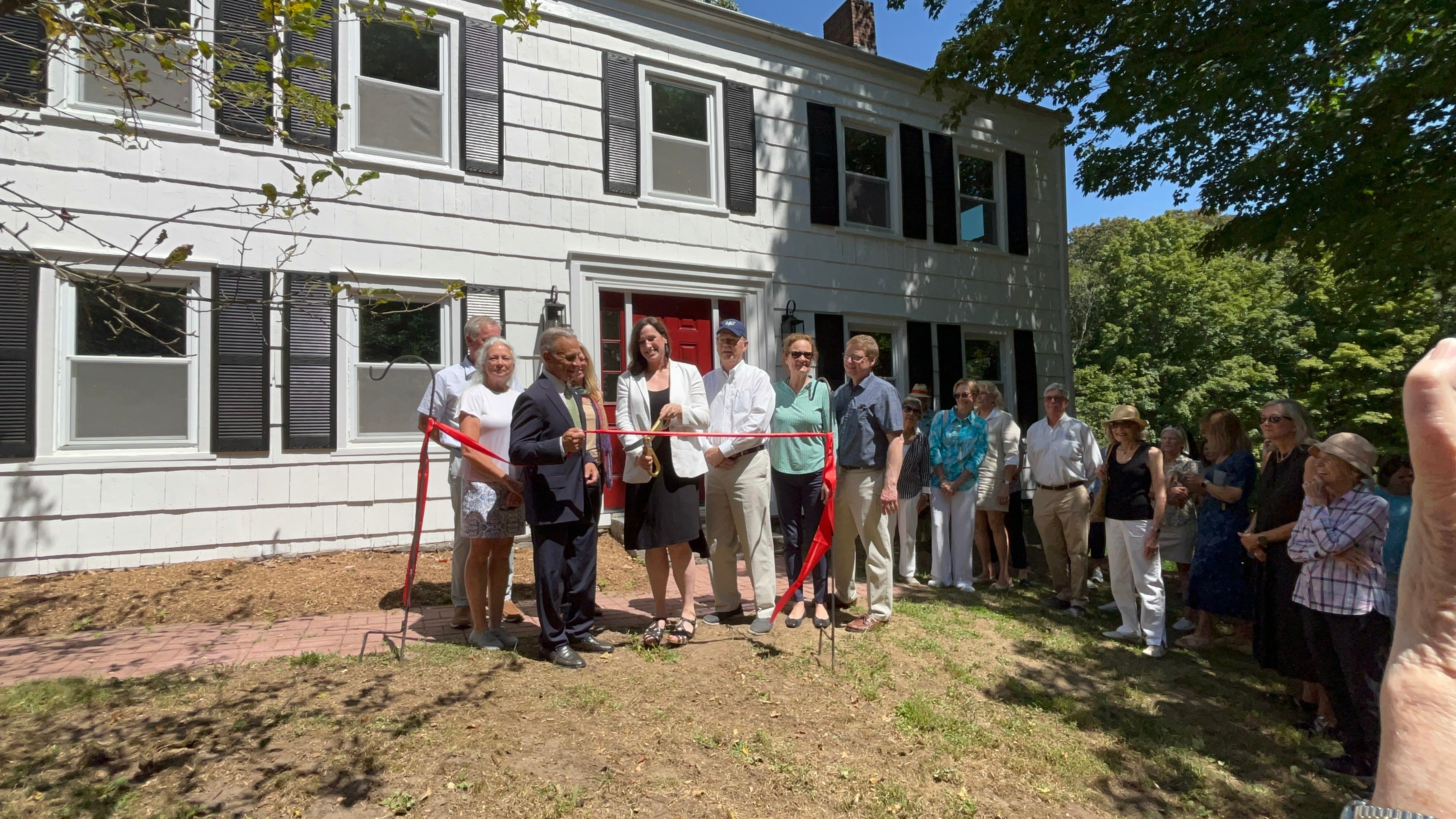 A group of people stand in front of the Mead House, cutting a red ribbon with a large pair of scissors.