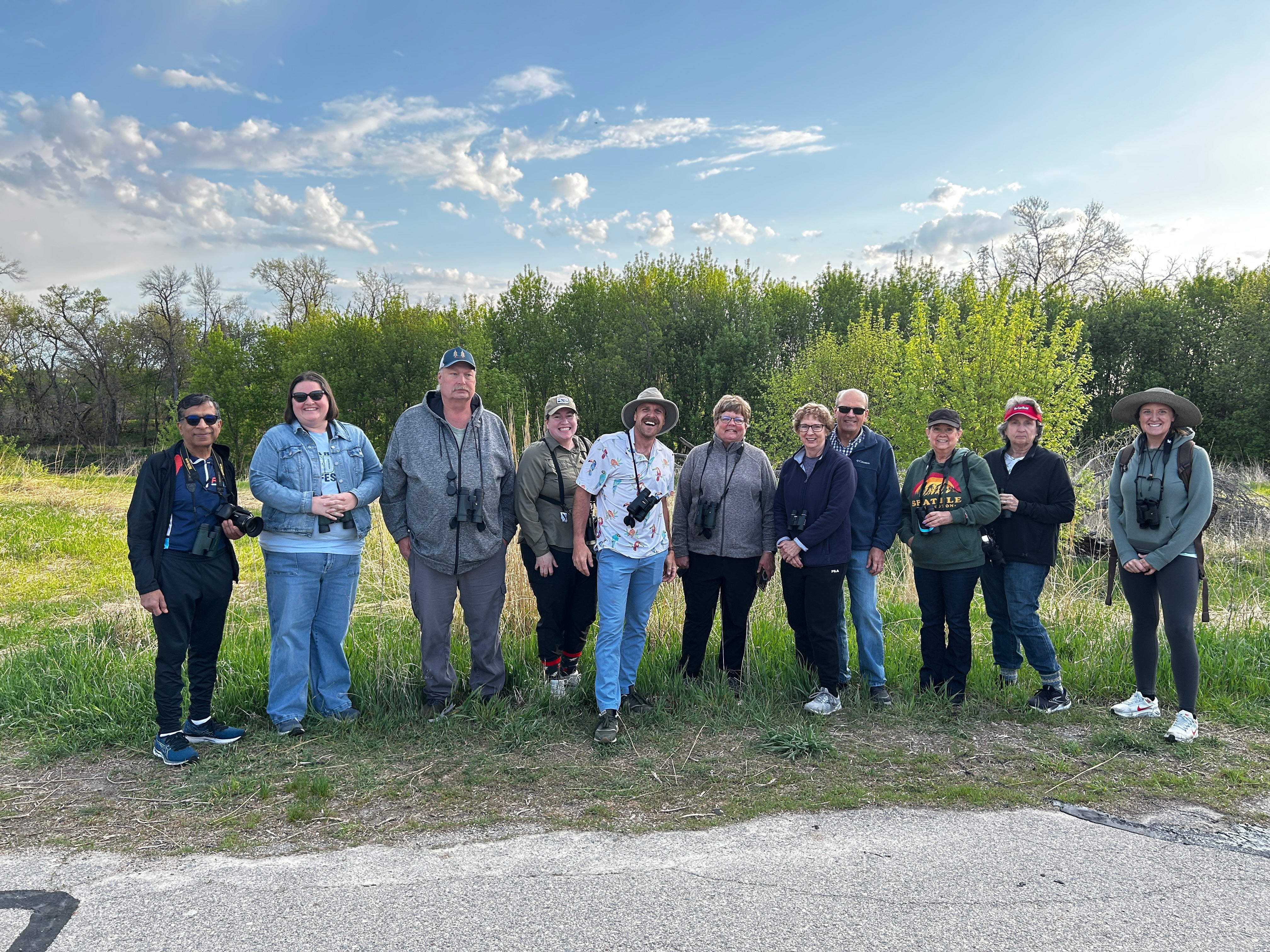 Group of birders outside on a sunny day with green trees and grass.