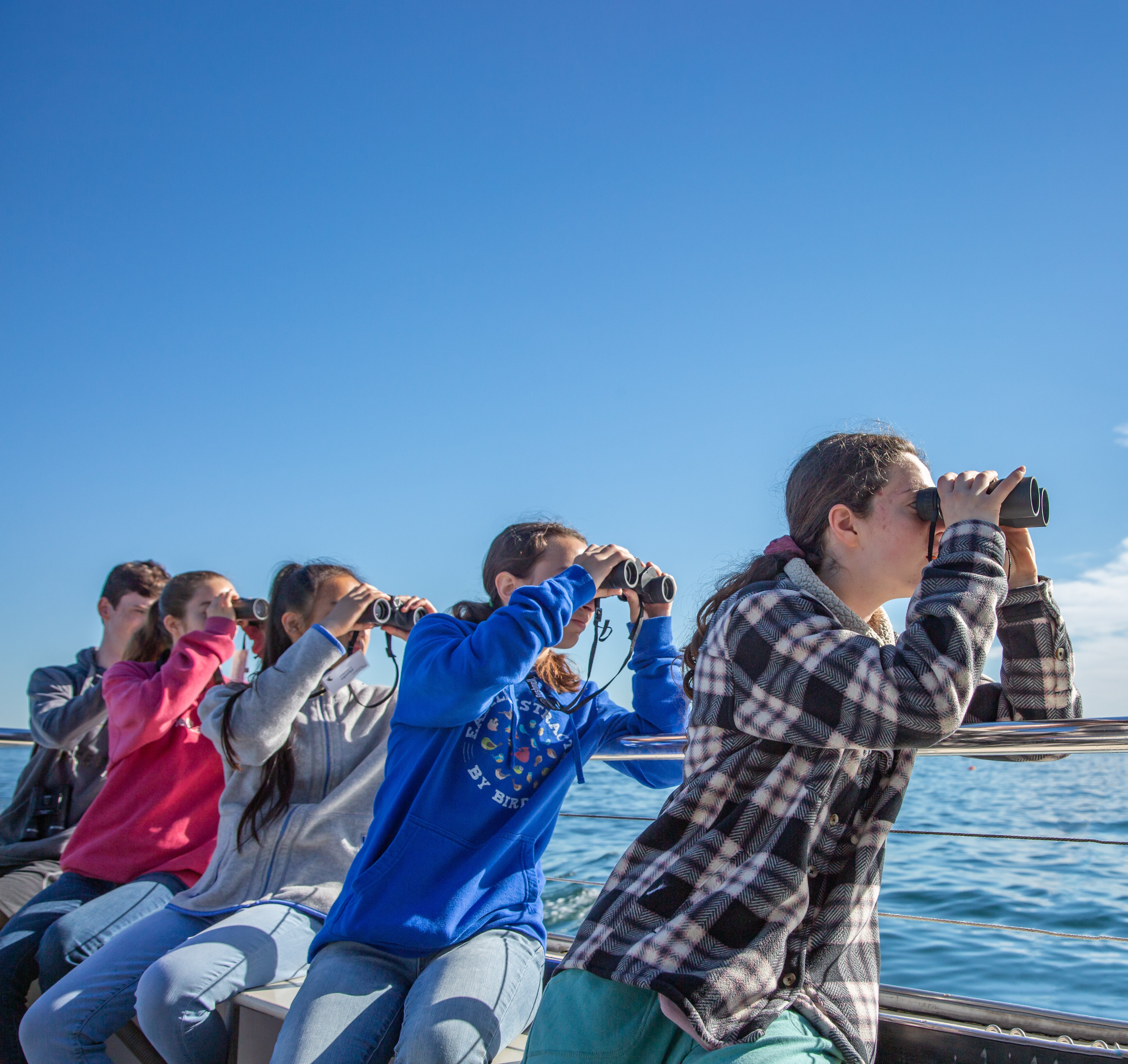 A group of teens looking through their binoculars while sitting on a boat.