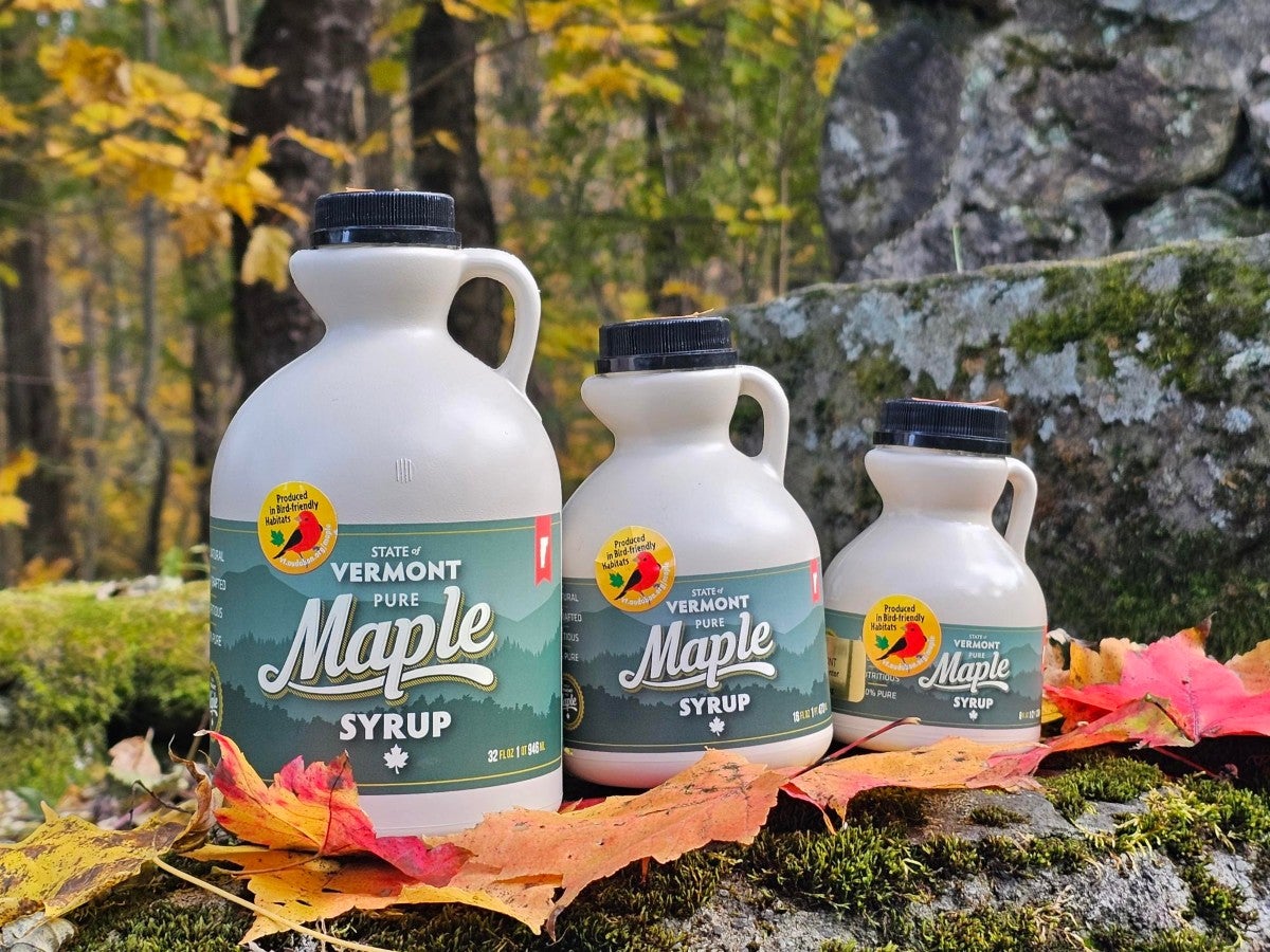 Three maple syrup bottles of varying sizes lined up from largest to smallest on a moss and leaf covered rock wall.