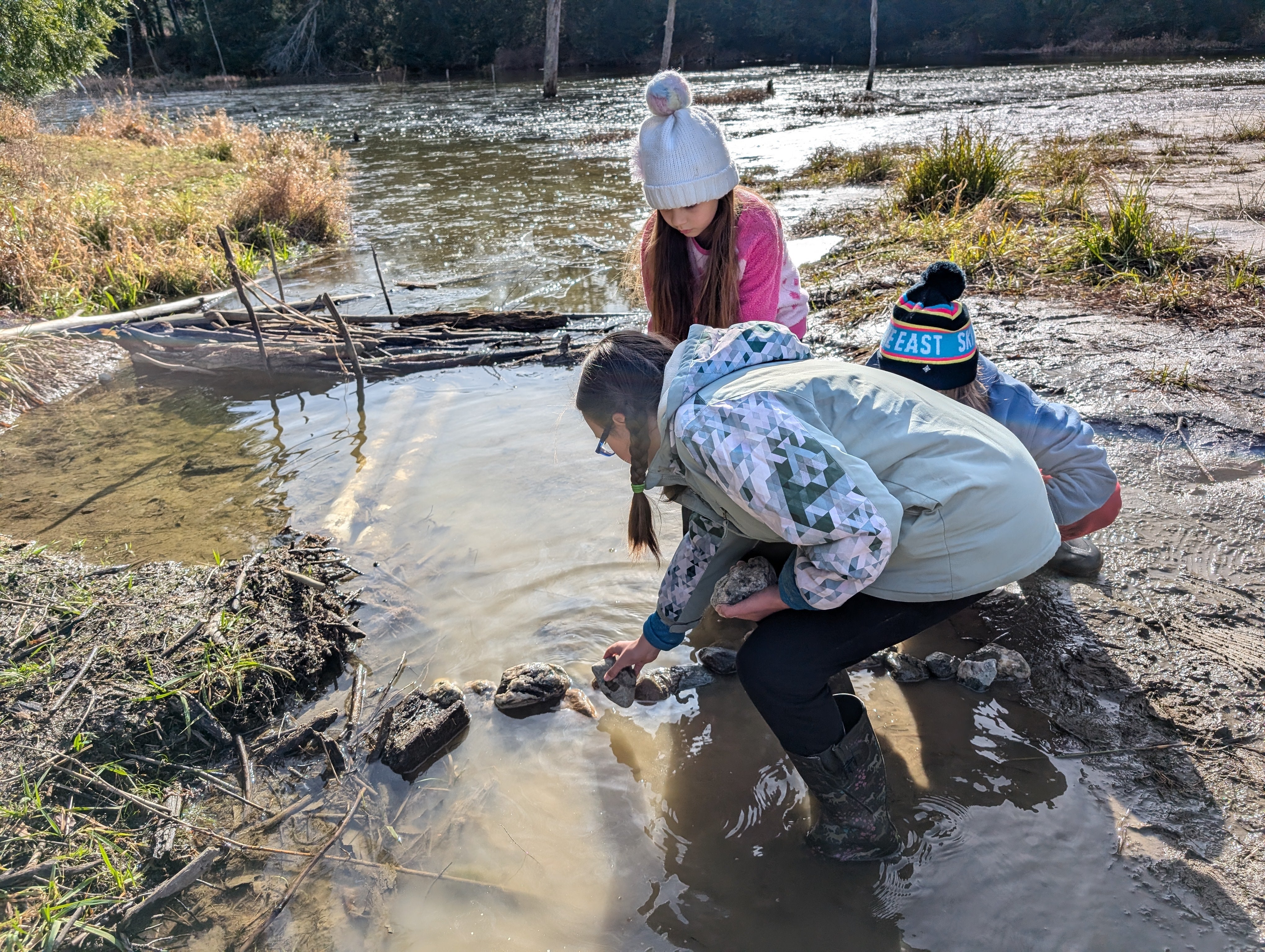 Three children crouch down in shallow water to investigate a beaver dam.