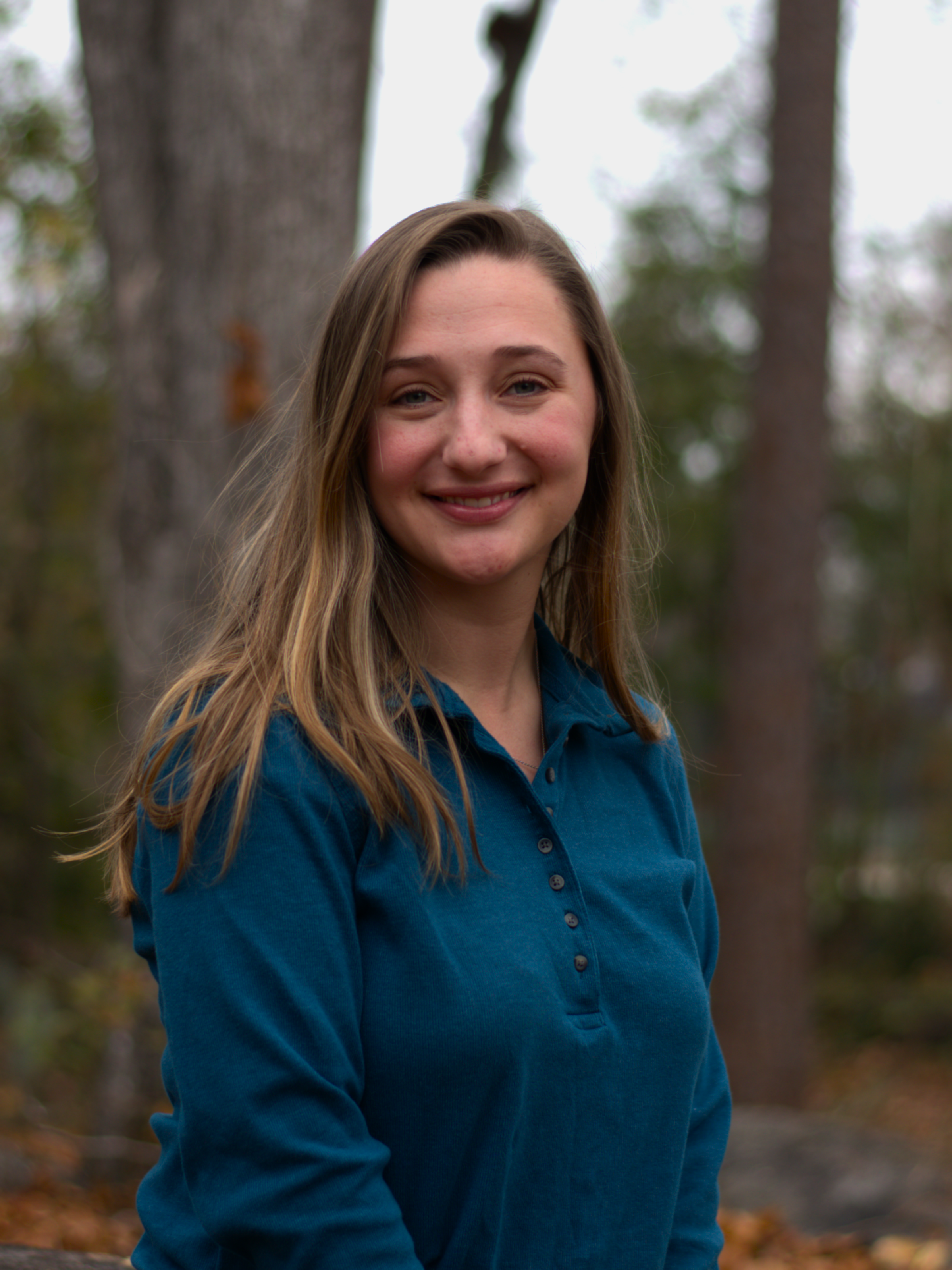 A smiling young woman with Blond hair in a forest wearing a blue shirt