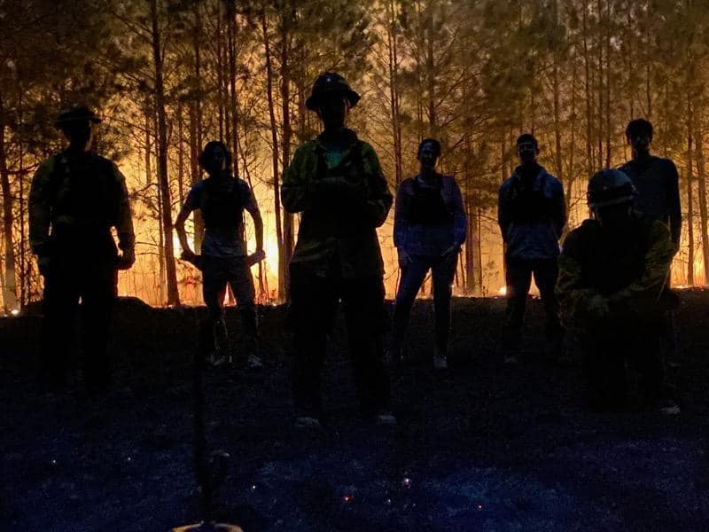 Silhouettes of the fire crew stand in front of a pine forest that in engulfed in flames at night
