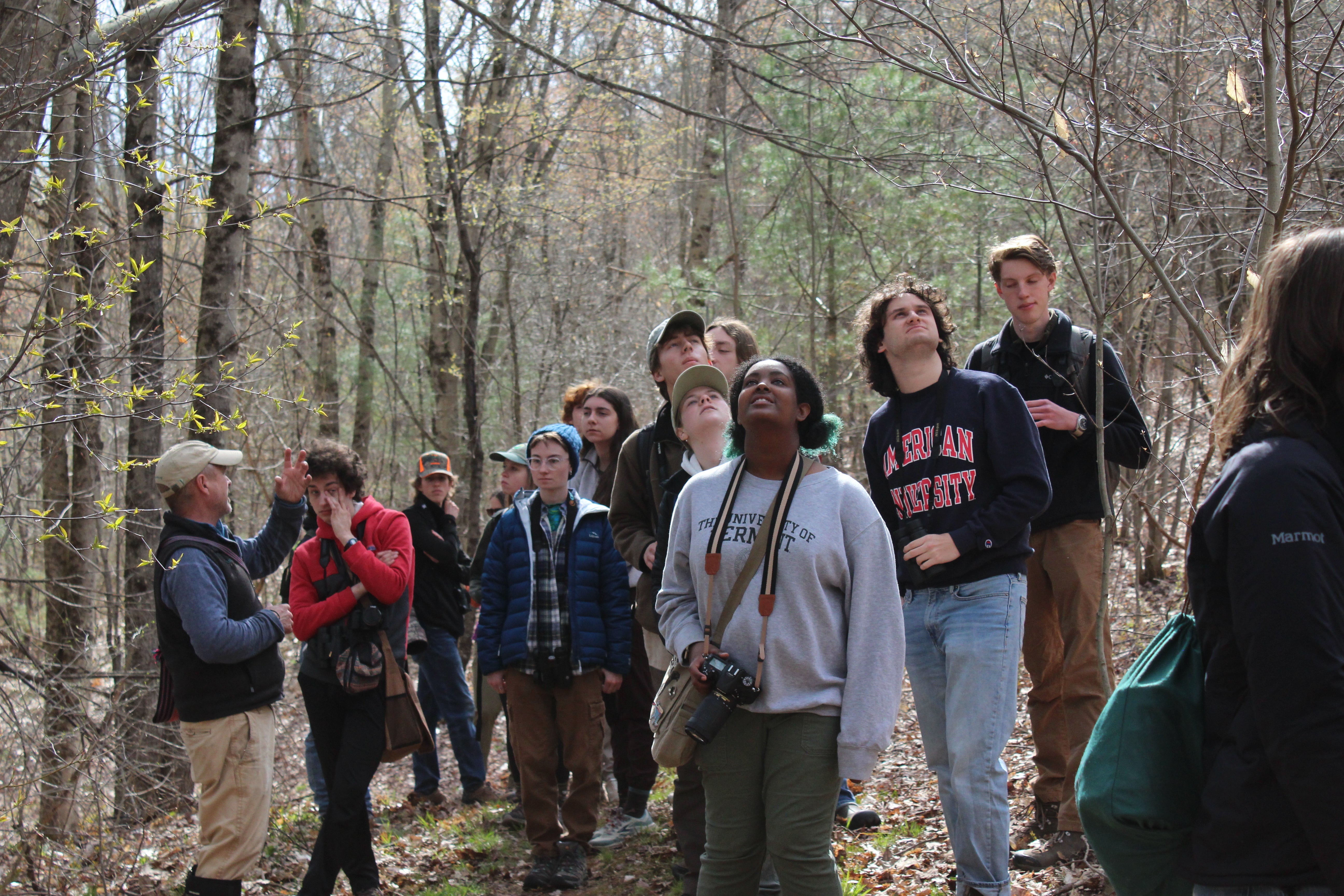 A group of college students gathered along a forested path looking up at the trees as �ԹϺ��� staff member speaks.