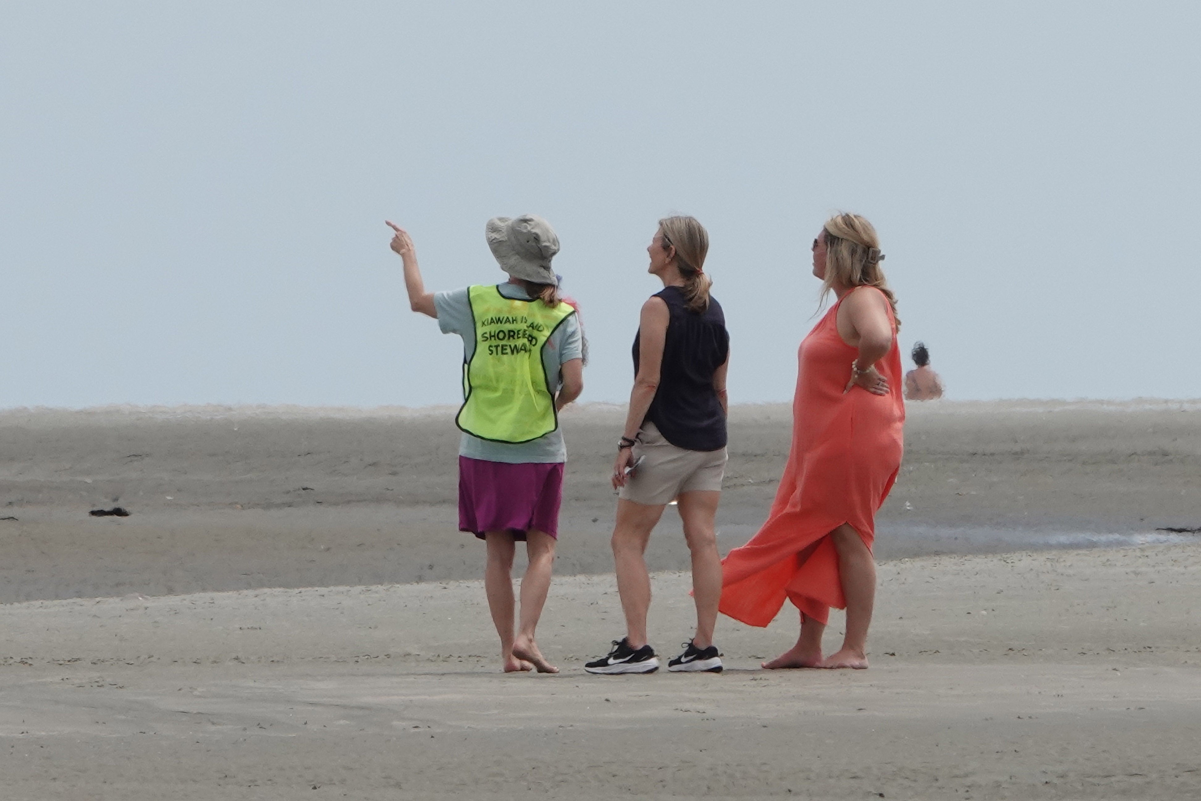 Two people stand on a beach and talk to a person in a bright green vest