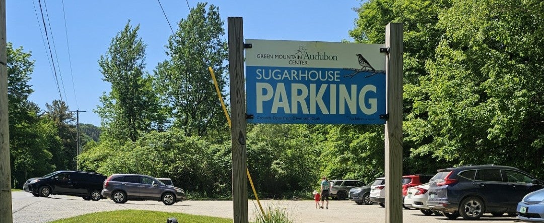 A photo of a gravel parking lot full of cars. A tall sign reads "Sugarhouse Parking".