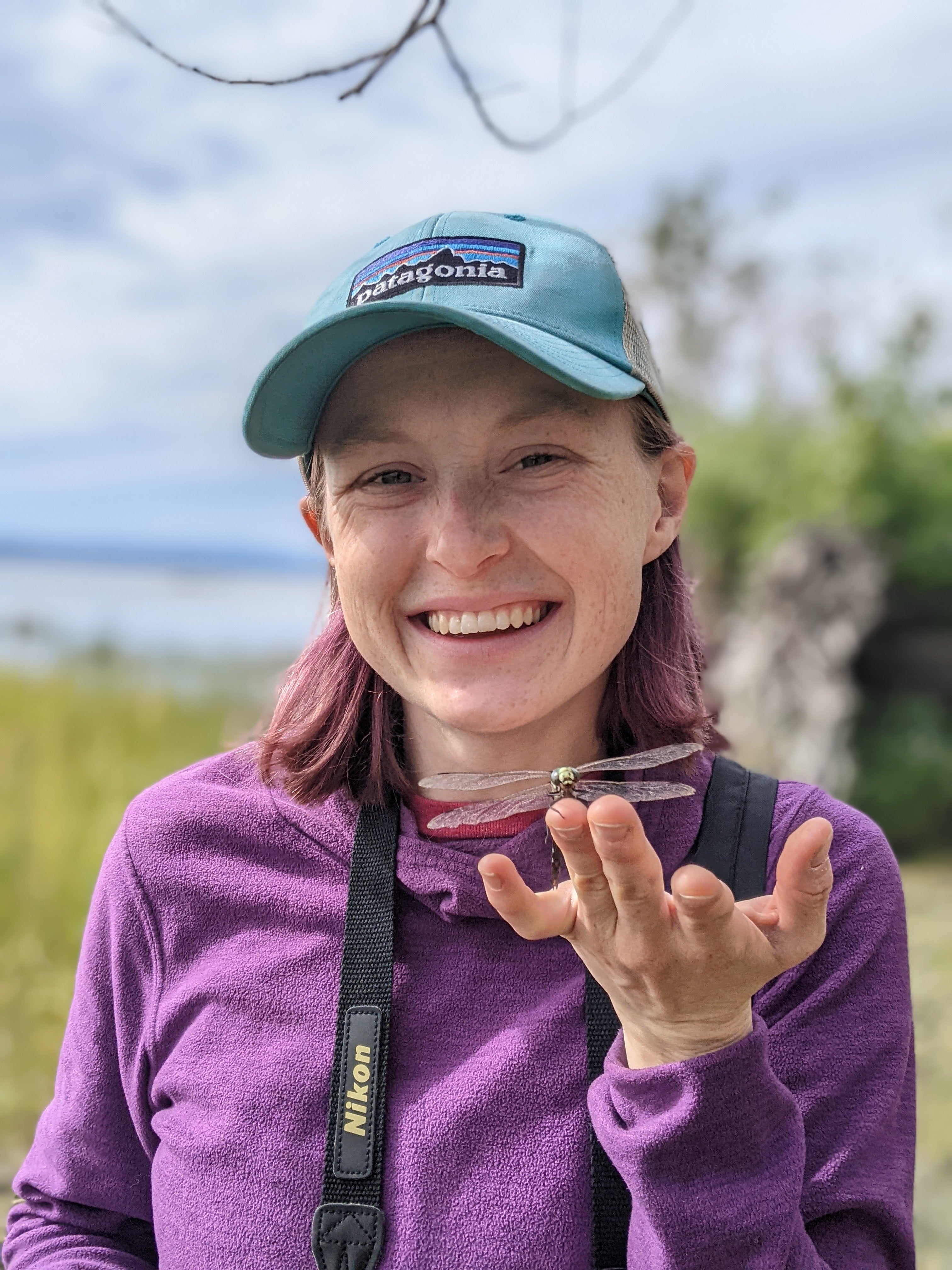 A photo of Emily Calder smiling with a dragonfly perched on her outstretched hand.