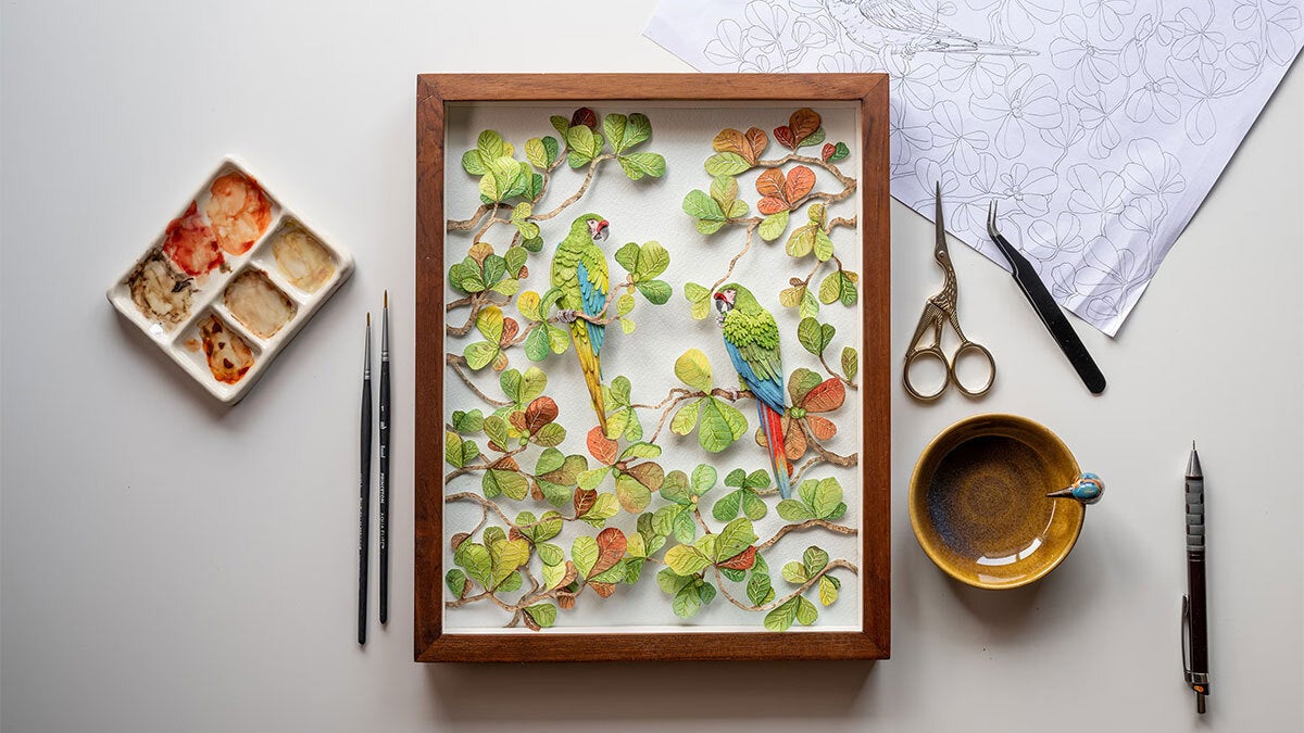 A framed paper artwork shows two great green macaws perched on branches. The birds and branches are crafted entirely from layered paper. Around the frame lie various art tools—scissors, paintbrushes, and tweezers.
