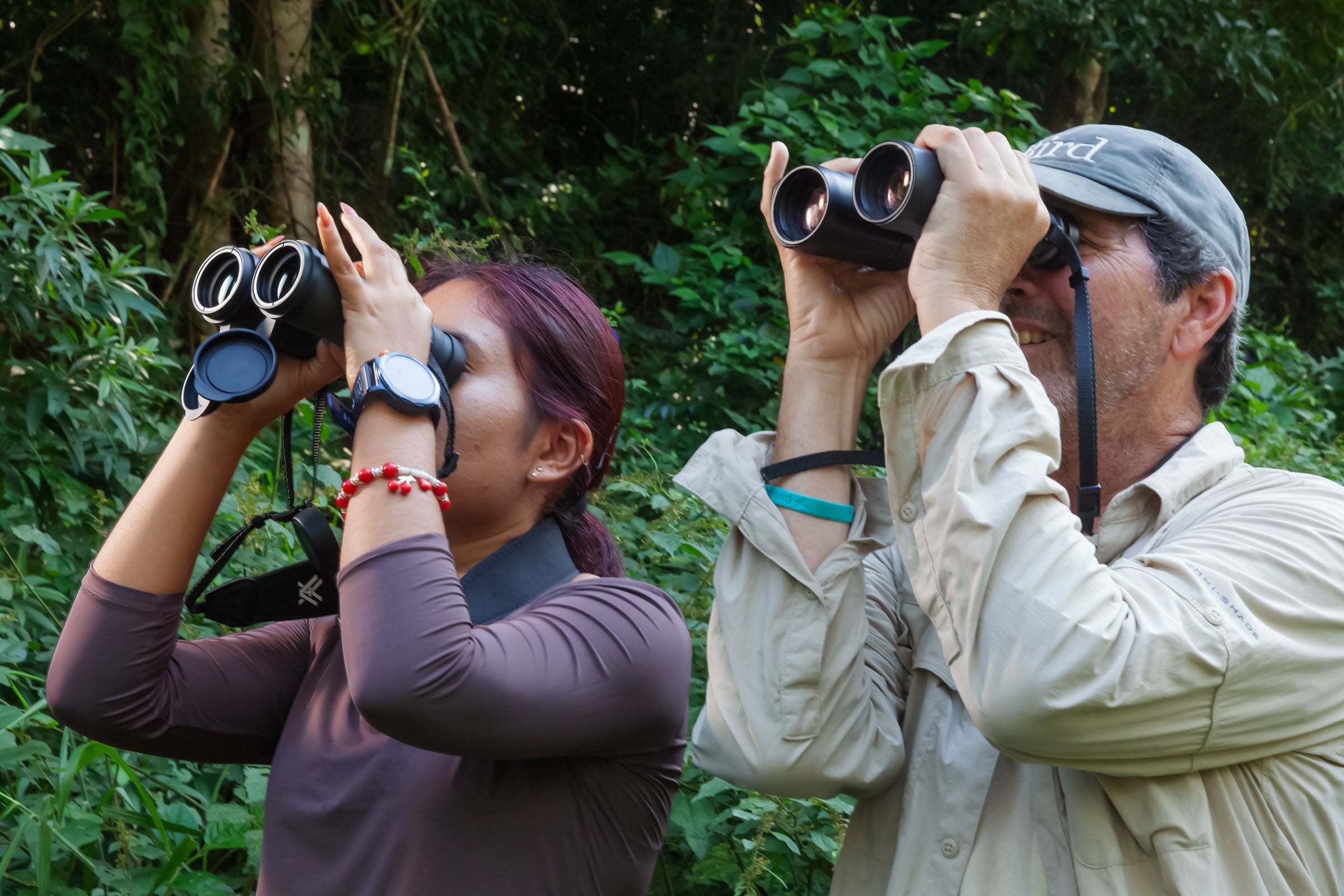 a woman and a man holding binoculars up to their eyes in a forest.