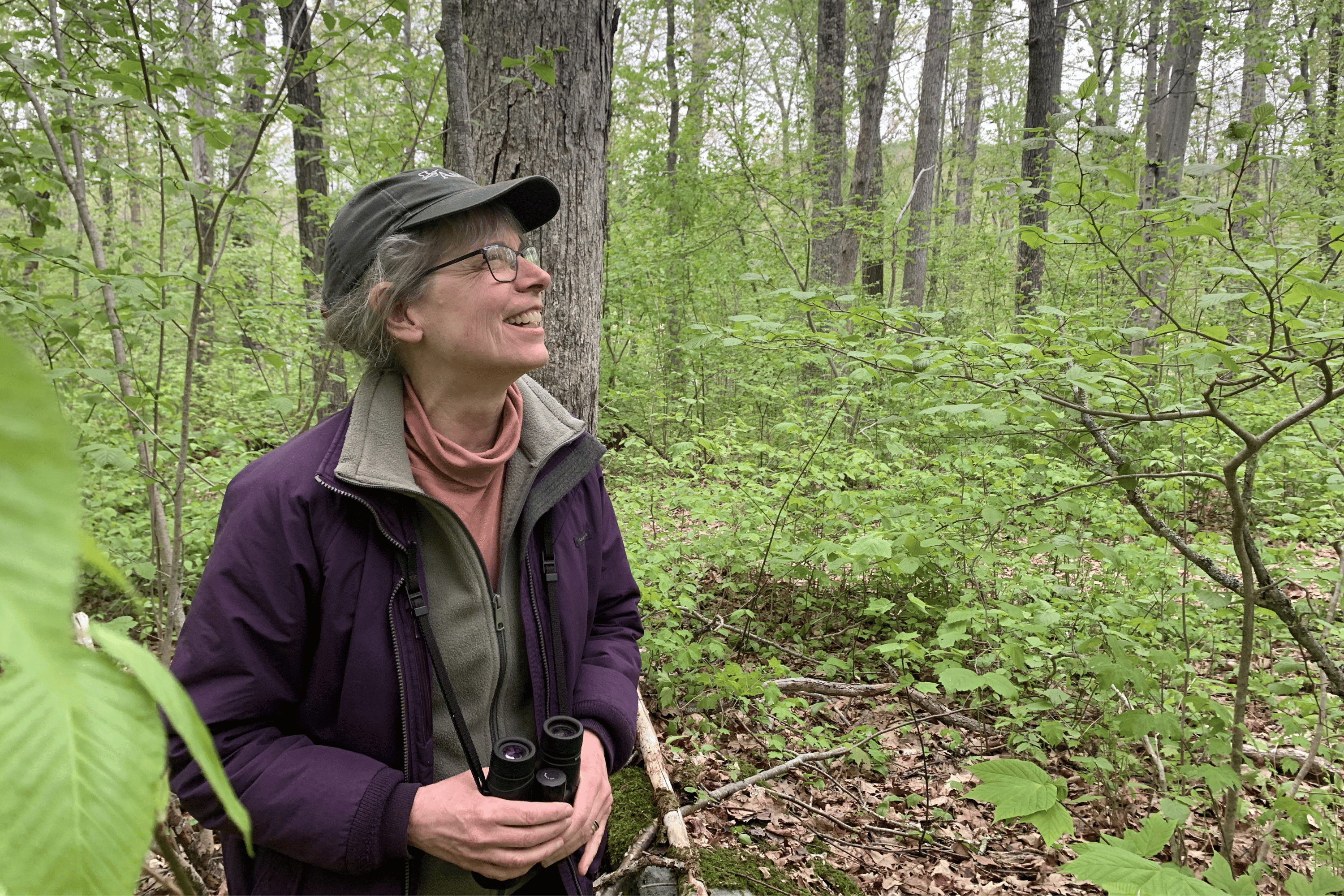 Eileen Fielding in a forested area, smiling while holding a pair of binoculars.