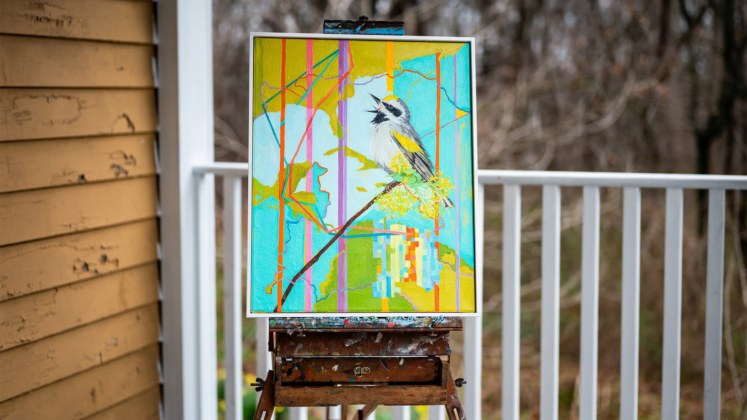 A painting of a Golden-winged Warbler with a colorful background sits on an easel on a porch.