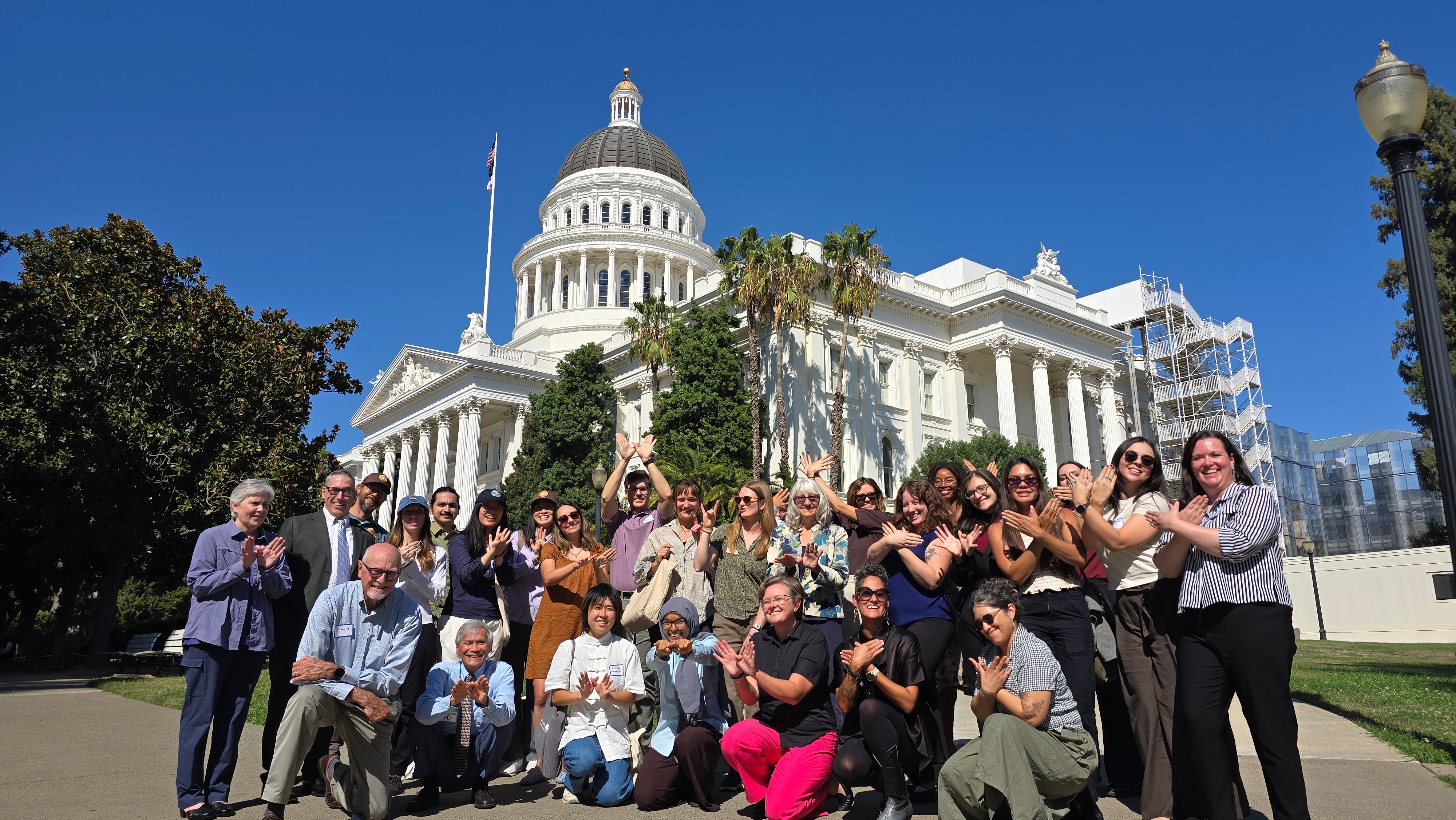 A diverse group of about 25 people pose in front of the California State Capitol building