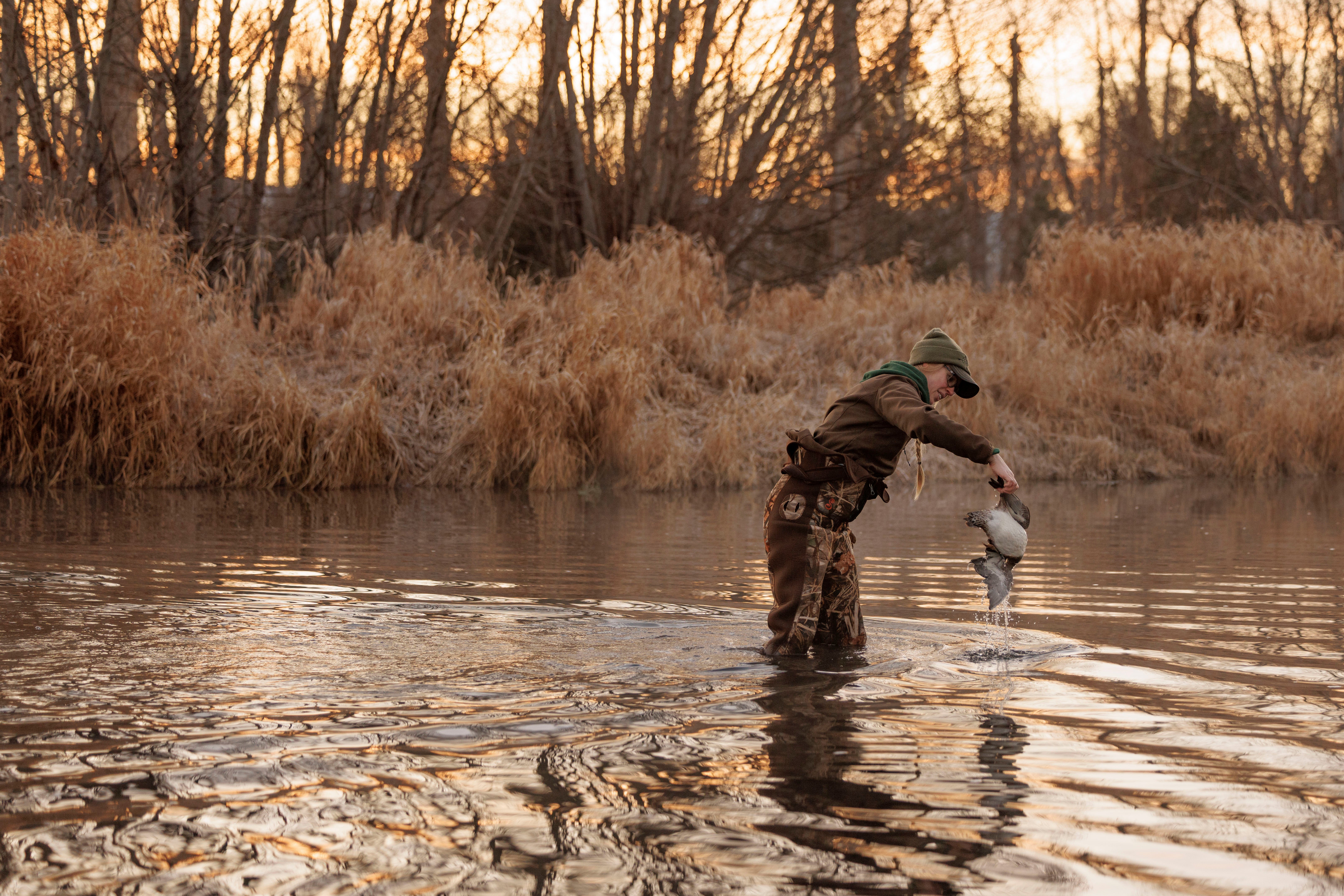 A person wearing waders standing in the water and holding a dead duck.