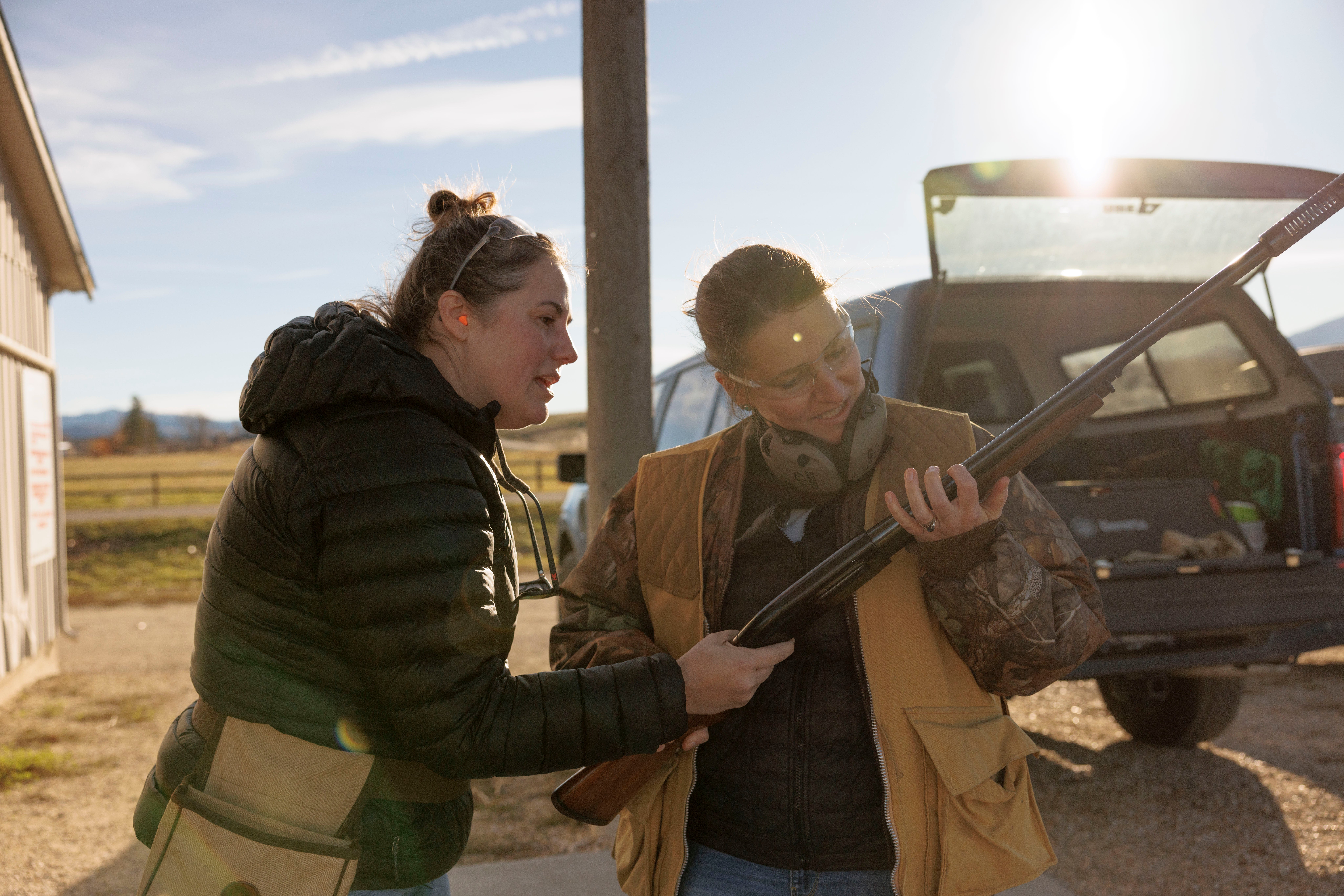 A person looking at a rifle held by another person, with a car and its open trunk in the background.