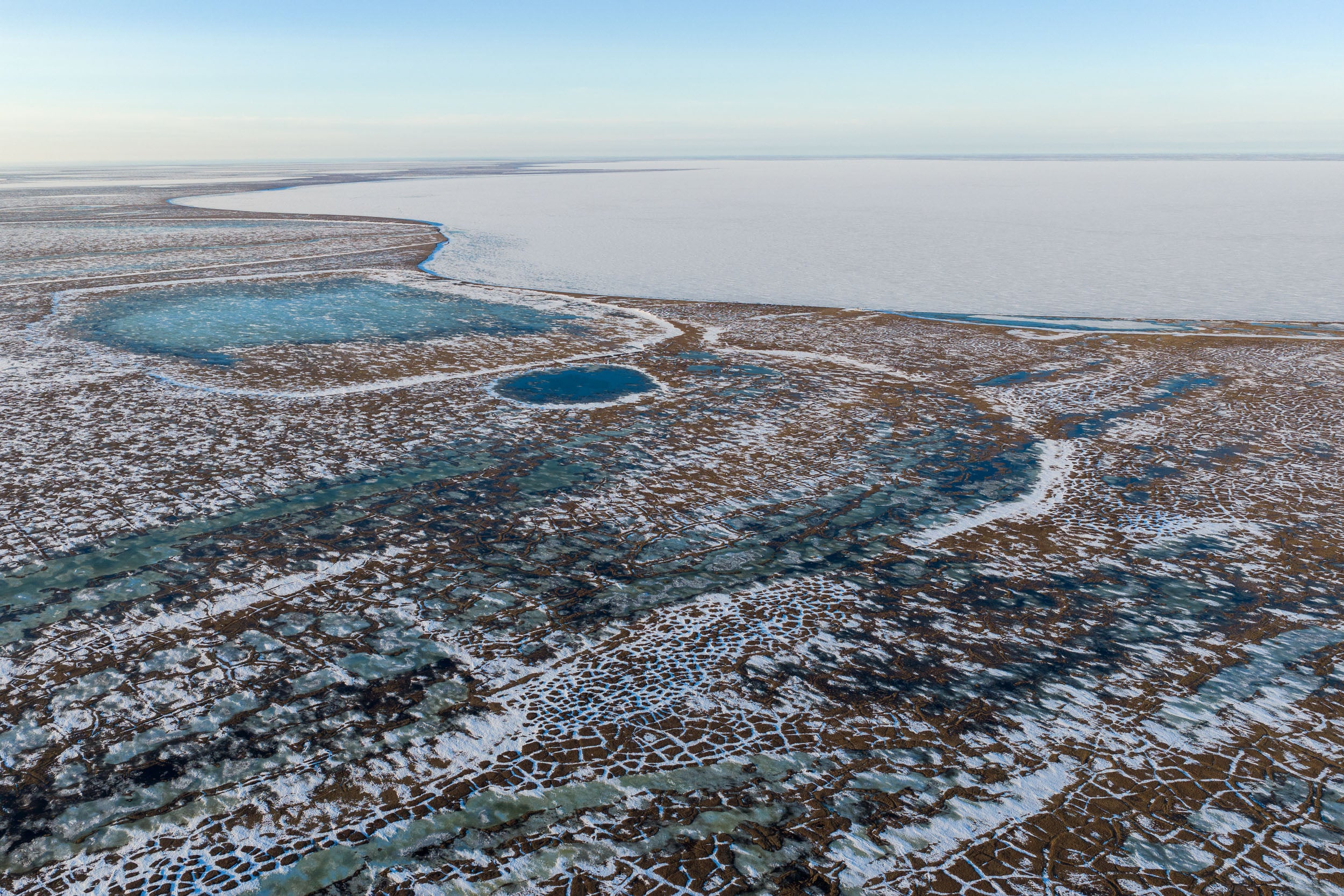 Aerial of thawing tundra on the side of a frozen lake. 