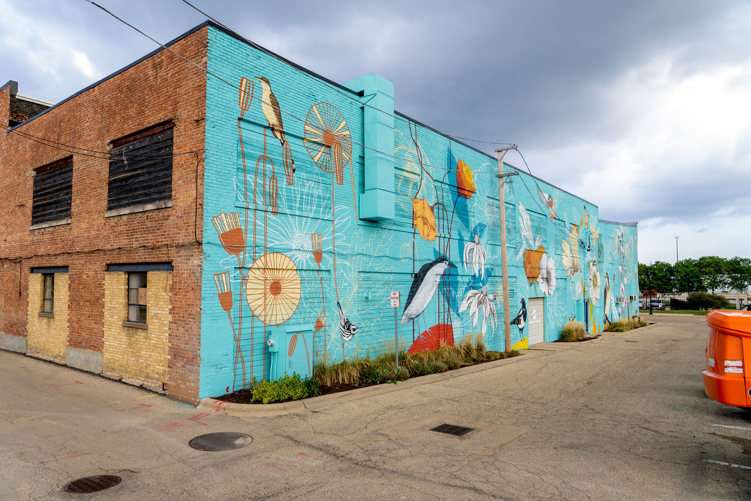 One side of an industrial brick building is painted turquoise blue and depicts birds perched on tall, elegant flowers. The mural by artist Yulia Avgustinovich includes bird species ranging from long-legged wading birds, like the Black-crowned Night Heron, to small songbirds, like the Black-and-white Warbler.