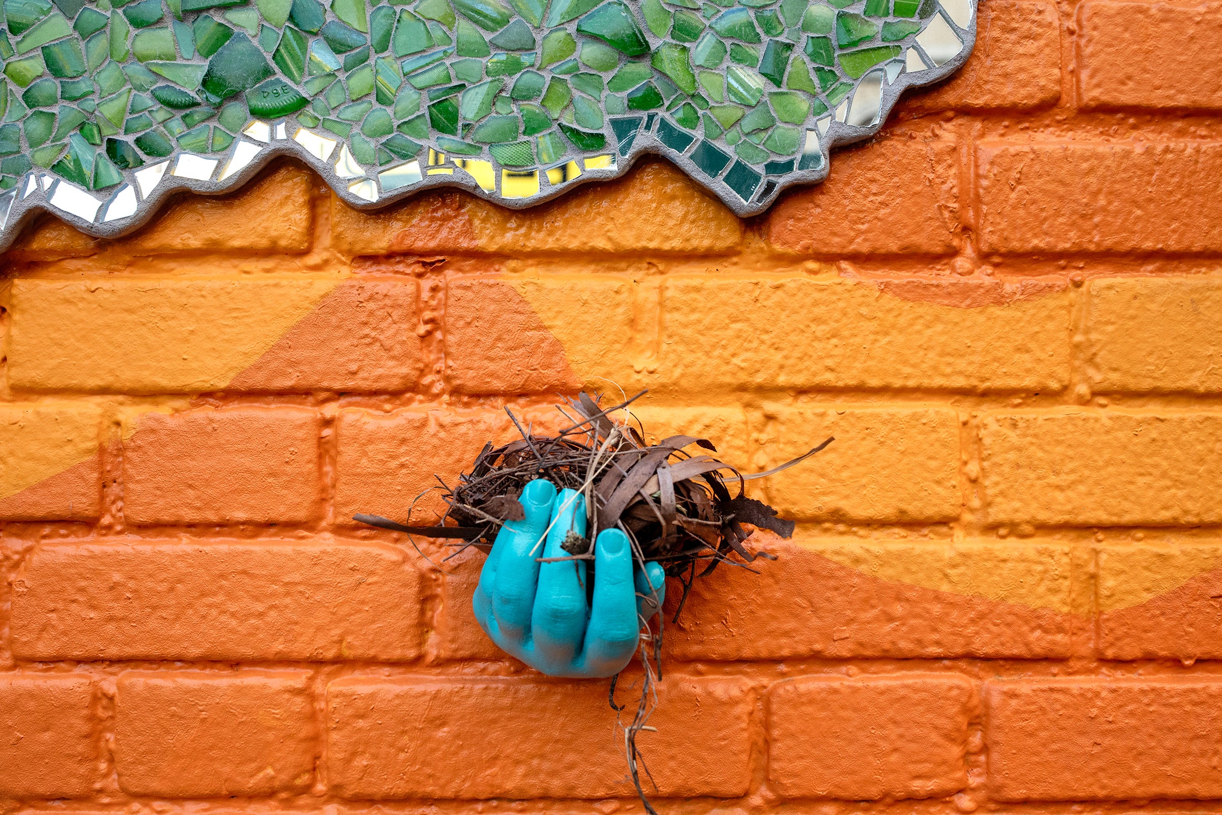 A concrete hand, painted robin's egg blue, juts out from a bright orange brick wall. Inside the hand is a small pile of brown twigs and bark resembling a birds nest. In the upper left quadrant, green and mirrored mosaic are visible.