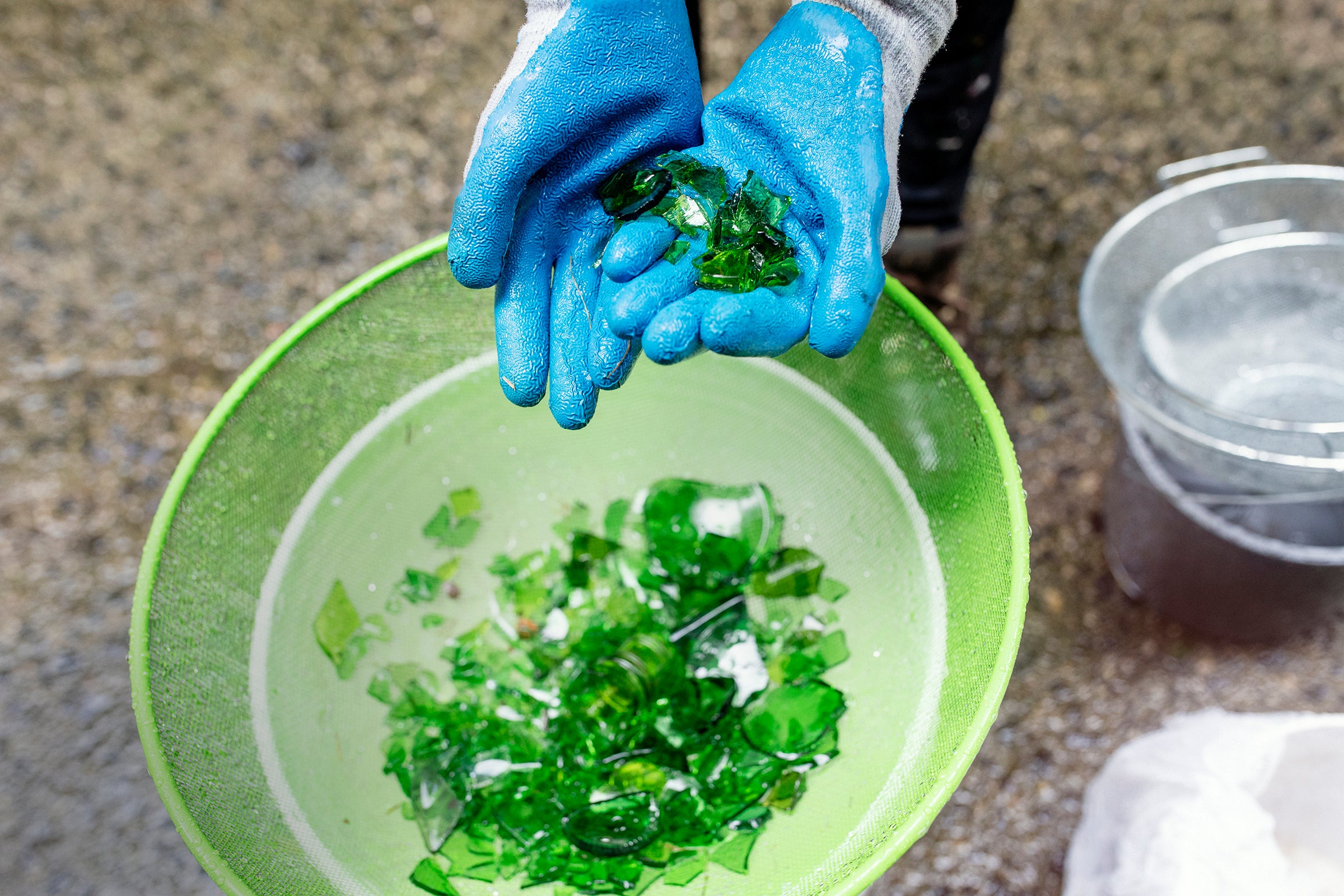 Hands wearing bright blue gardening gloves cup shards of emerald green glass over a lighter green pan.