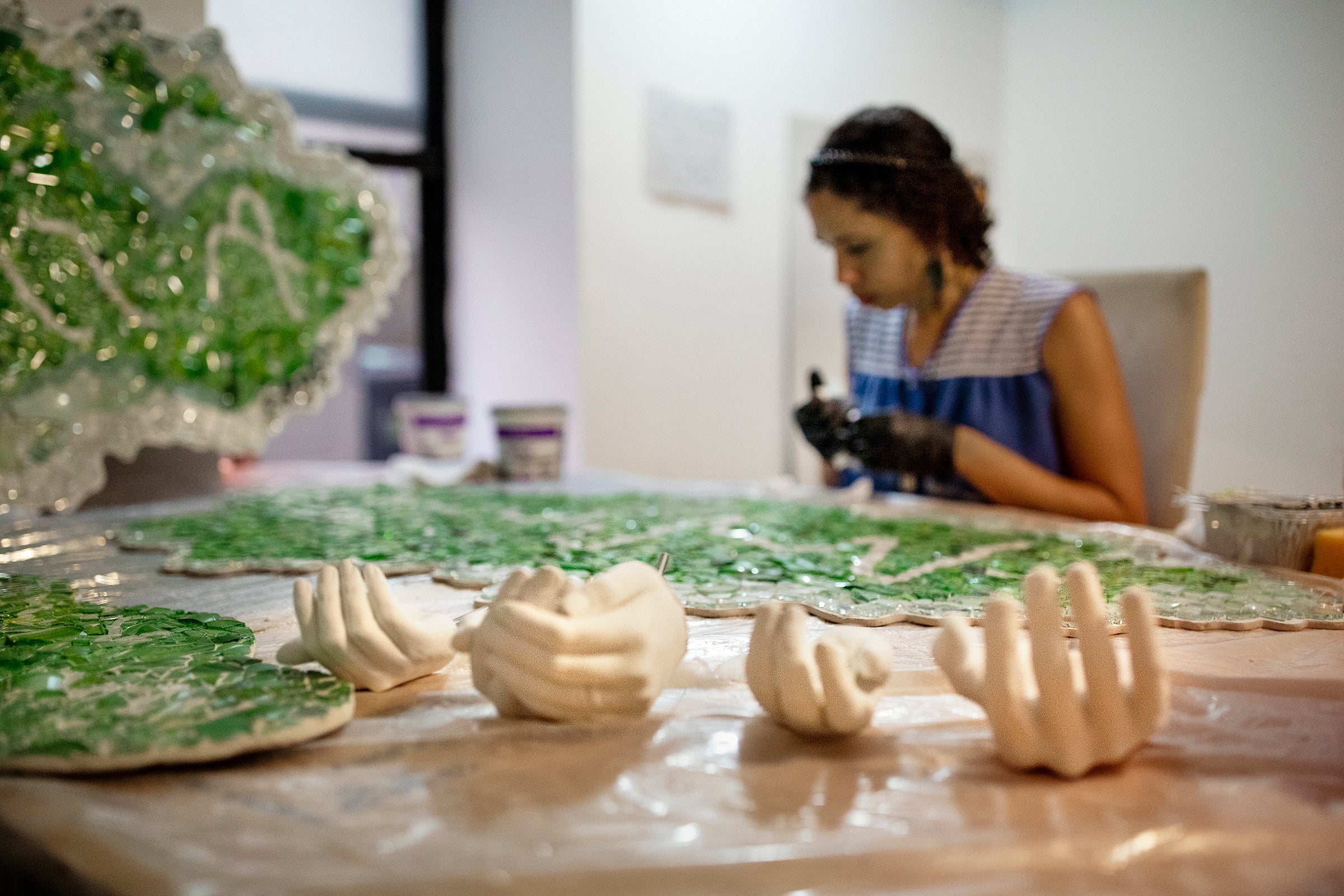 A woman sits in concentration at a long table in a white-walled room. In the foreground are sections of green and clear glass mosaic clouds and four hands cast from concrete.