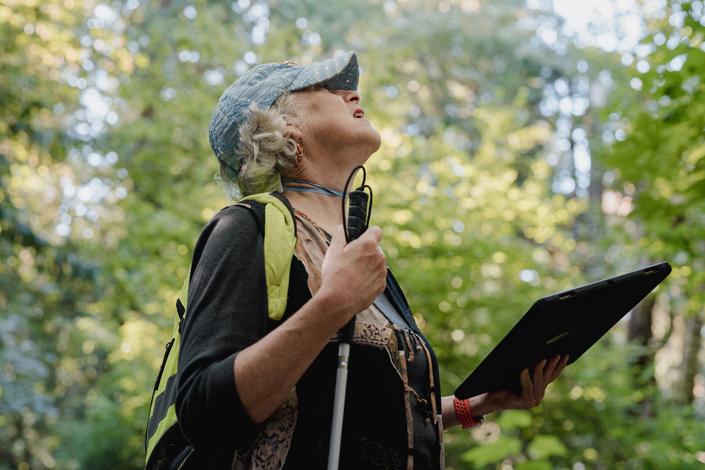 A woman holding an iPad and a walking stick looks up into the trees in a forest.