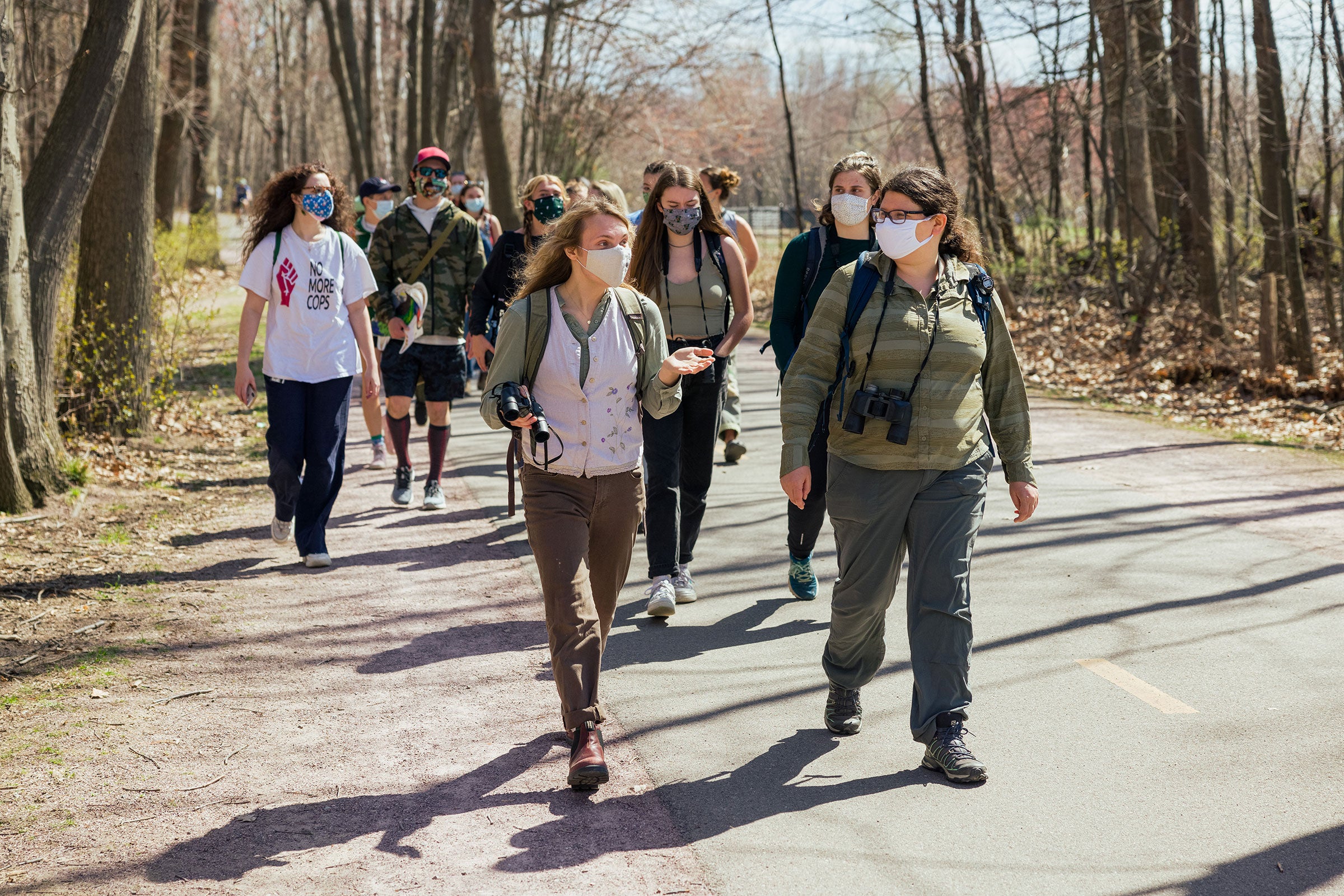 Around one dozen University of Vermont college students wearing face masks and binoculars around their necks walk down a road lined with brown trees during an outdoor class in Spring 2021.