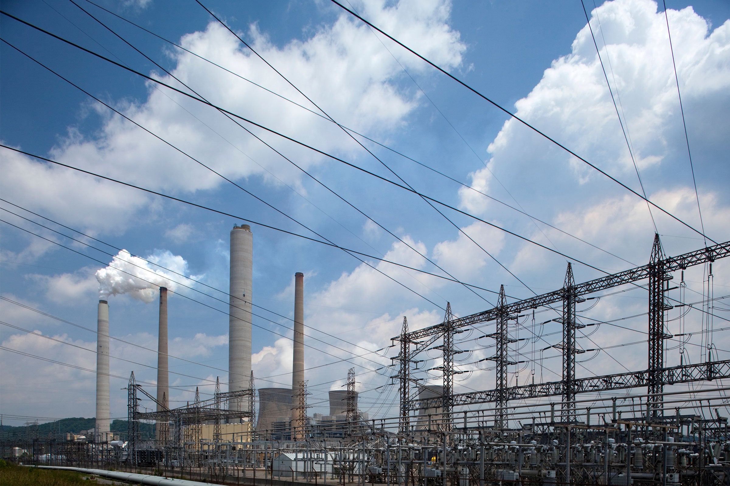 Powerlines criss-cross a blue sky; in the distance smokestacks from a coal-fired power plant emit smoke.