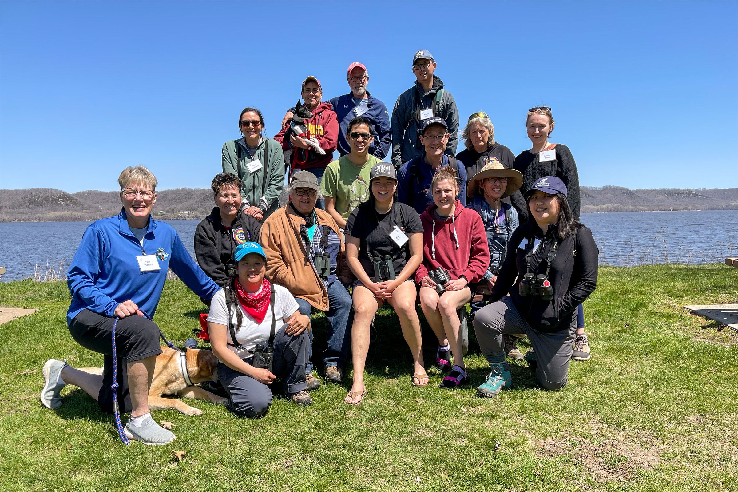 A group of about 15 people poses for a portrait by a lake. Many wear outdoor hats and gear, and name tags.