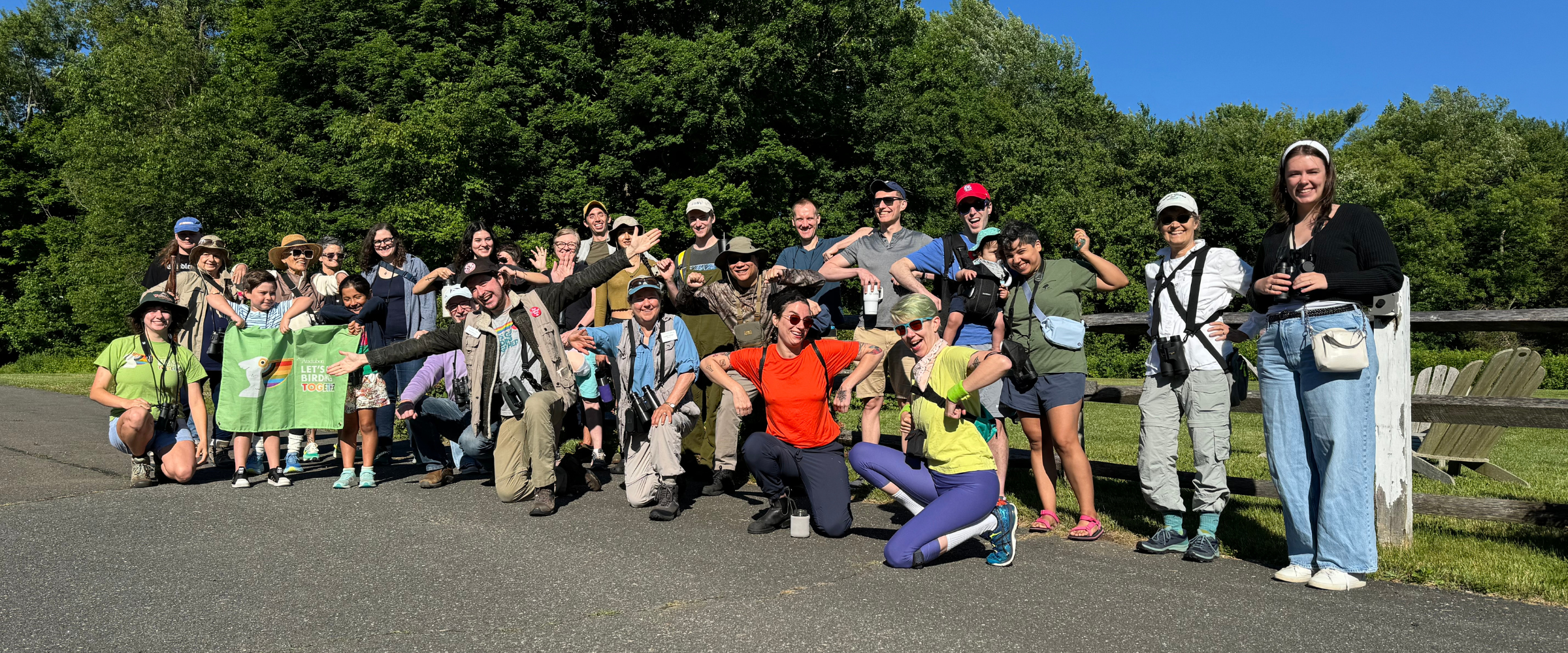 A large group of people with binoculars on a path at the Greenwich �ԹϺ��� Center, posing for a photo and smiling. One person is holding up a Let's Go Birding Together banner.