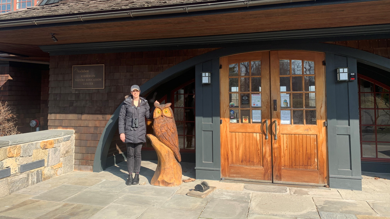 Rochelle Thomas stands in front of the Kimberlin Nature Education Center, posing next to a large wooden owl sculpture that is nearly her height.