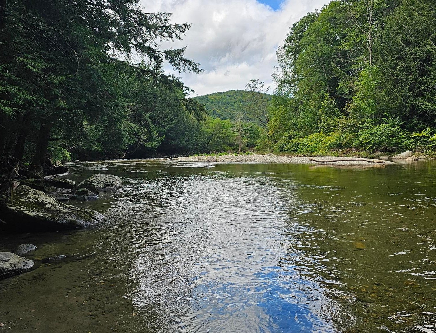 A landscape photo of a slow moving shallow section of the Huntington River. Framed by green trees. There is a small hill on the horizon.