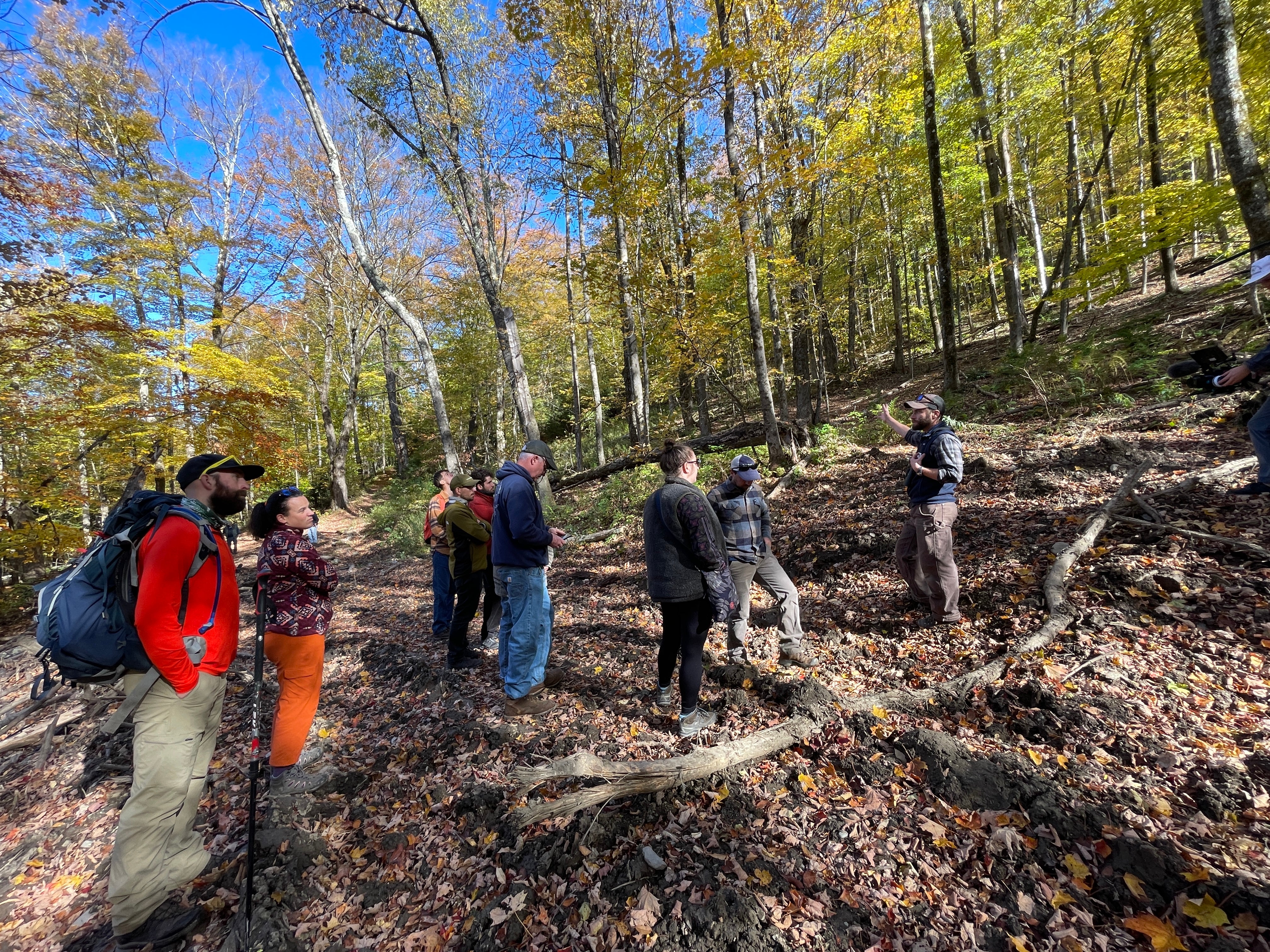A group of people gathered together in a circle on a forested trail in early fall. The leaves on the trees are just turning yellow.