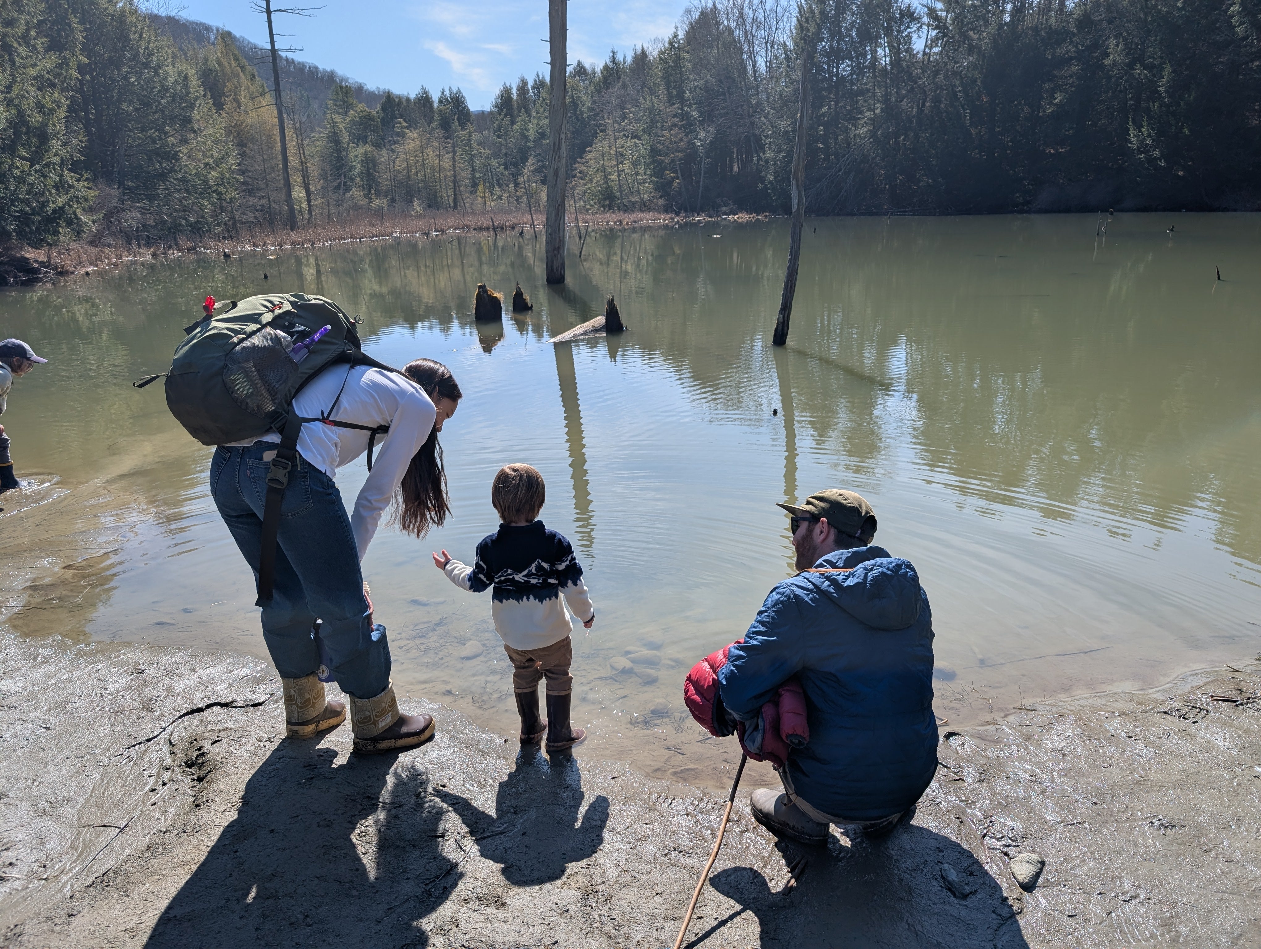 A kid stands by the edge of the pond with a teacher on one side and did dad on the other side crouching down and looking out at the pond