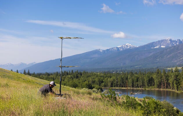 William Blake sits on the ground next to a Motus antenna, itself sitting in a green field that slopes into a river valley.