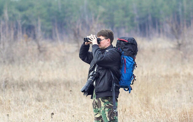 A man wearing camouflage pants and holding a camera and large backpack stands in a field and looks through binoculars.