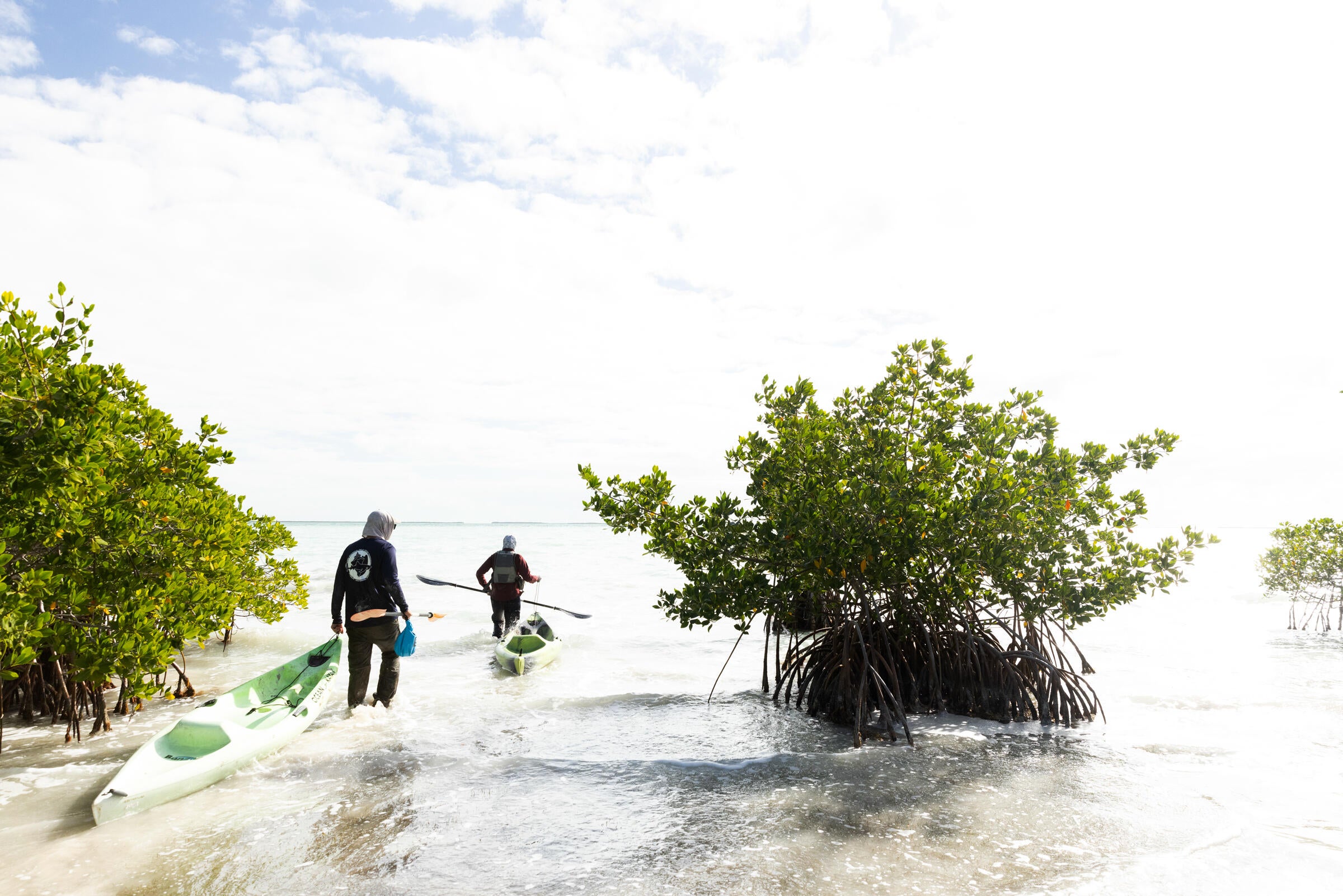 Two people wade through shin-high water each pulling a kayak behind them.