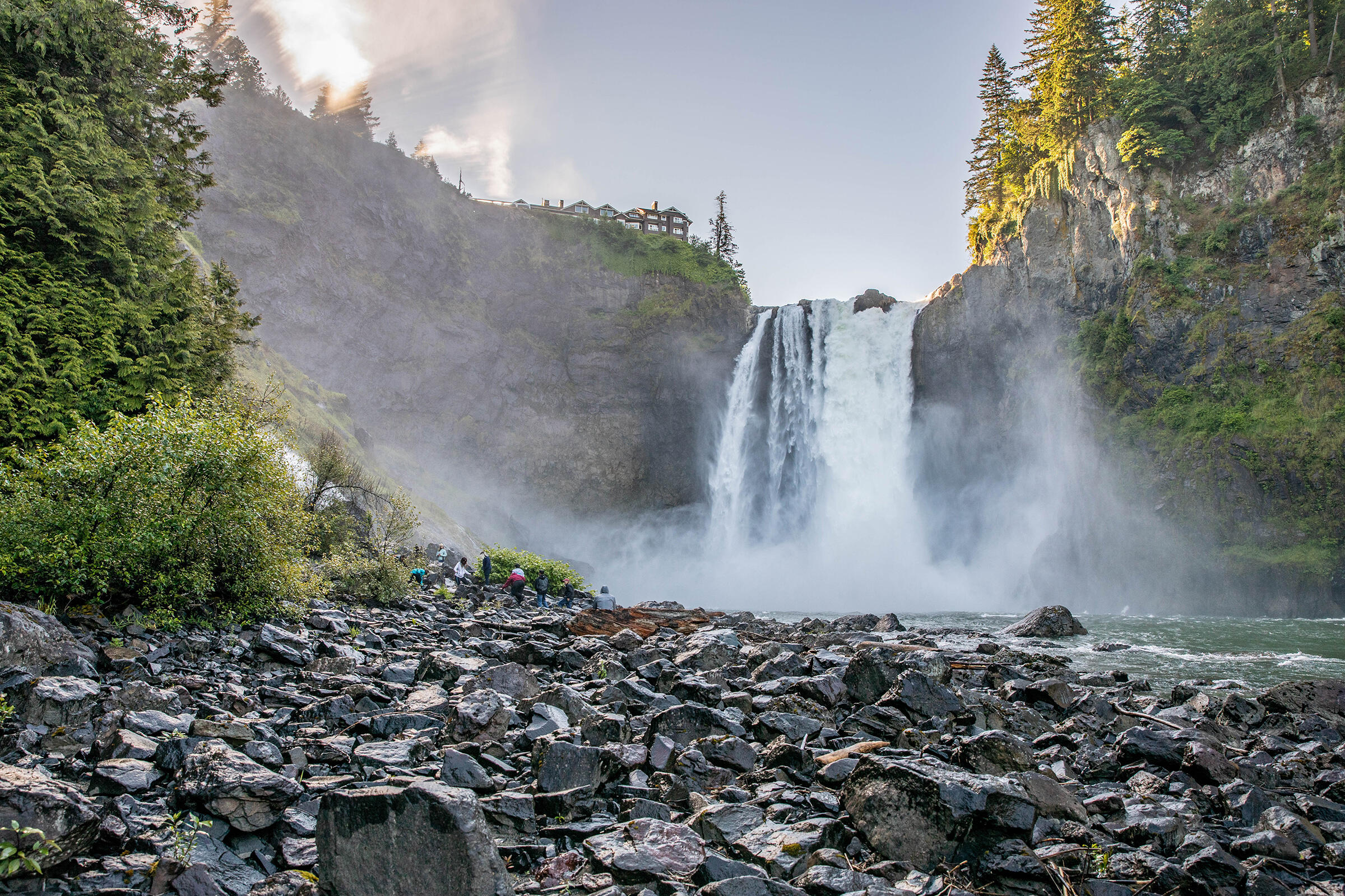 A massive waterfall pours over a cliff into the river below. On a rocky area on the shore, a small group of people gather in the distance. 