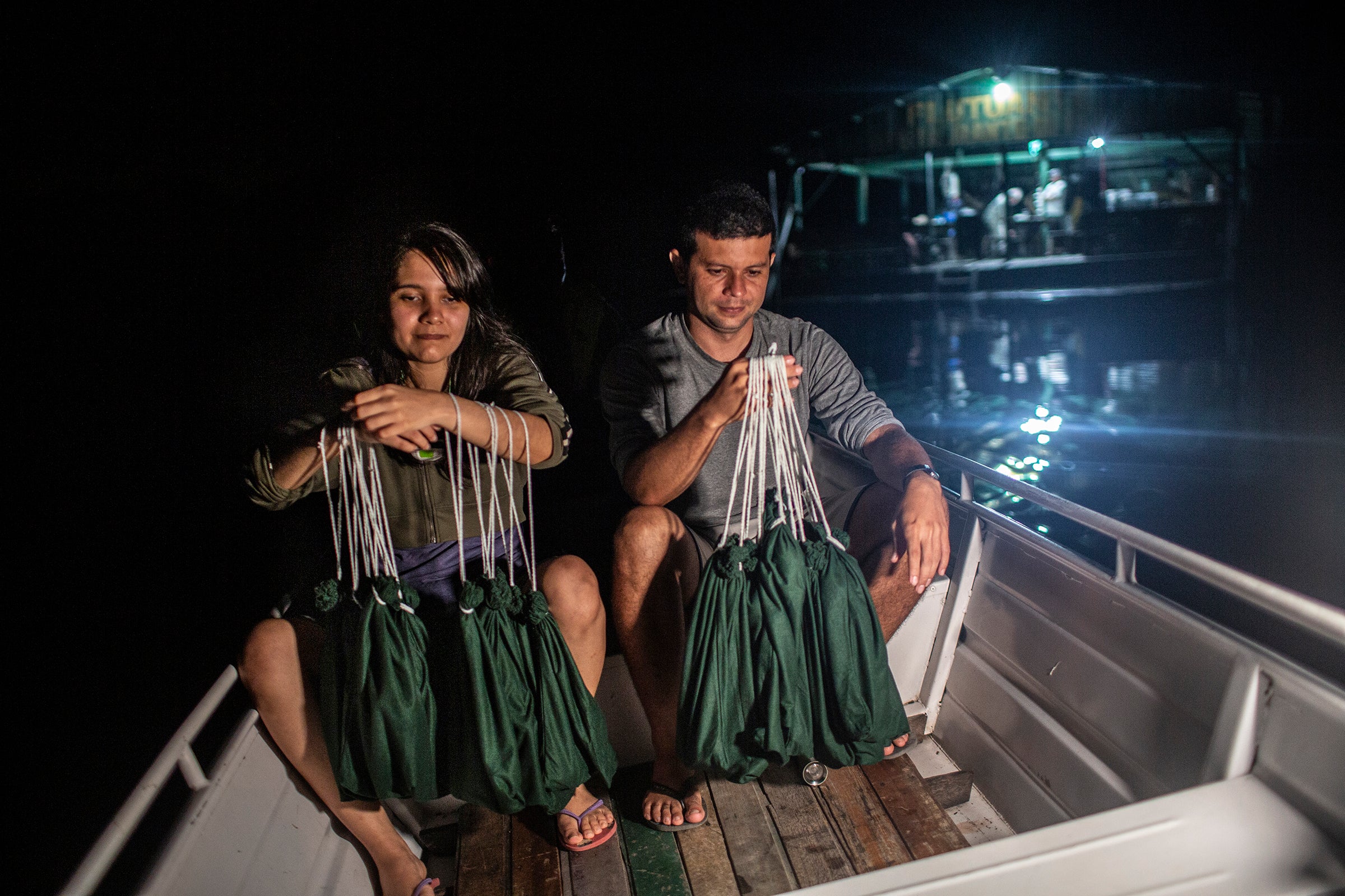 At night two people in small a boat hold many green bags. Behind them is a floating research structure.