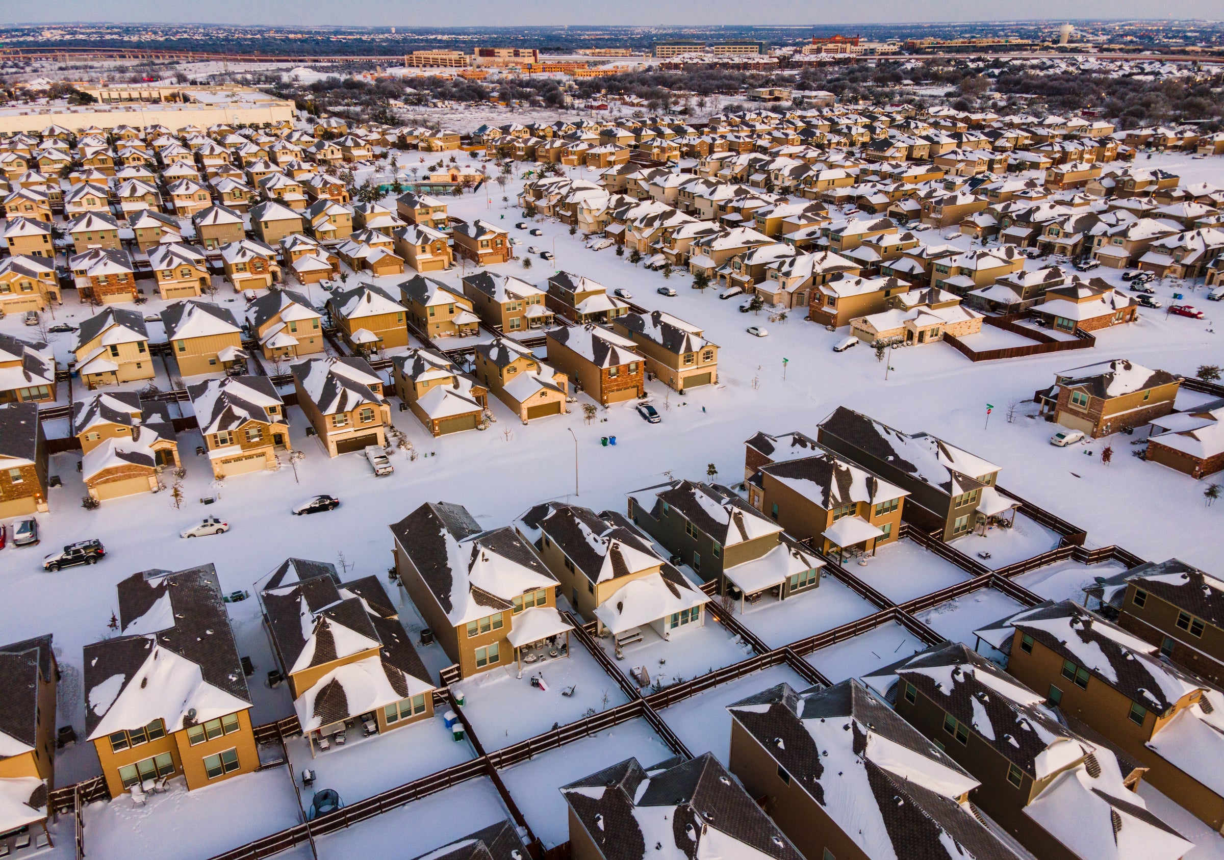 A blanket of snow coats the roads and roofs of rows and rows of houses. The neighborhood is seen in an aerial view. 