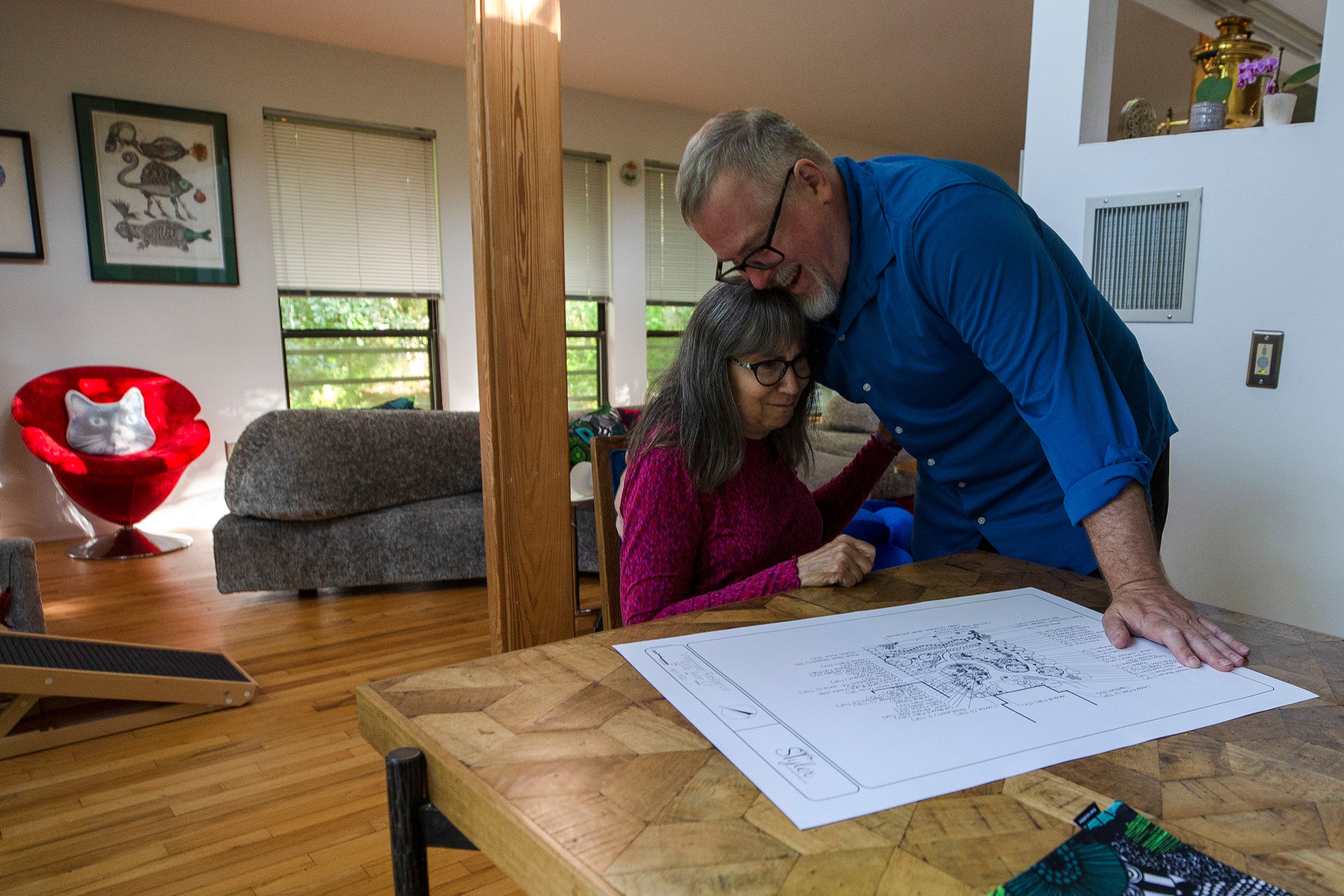 A man in a blue shirt hugs a women in a pink shirt who is sitting at a table, with blueprints spread out before her. In the background are a bright red chair and a gray couch.